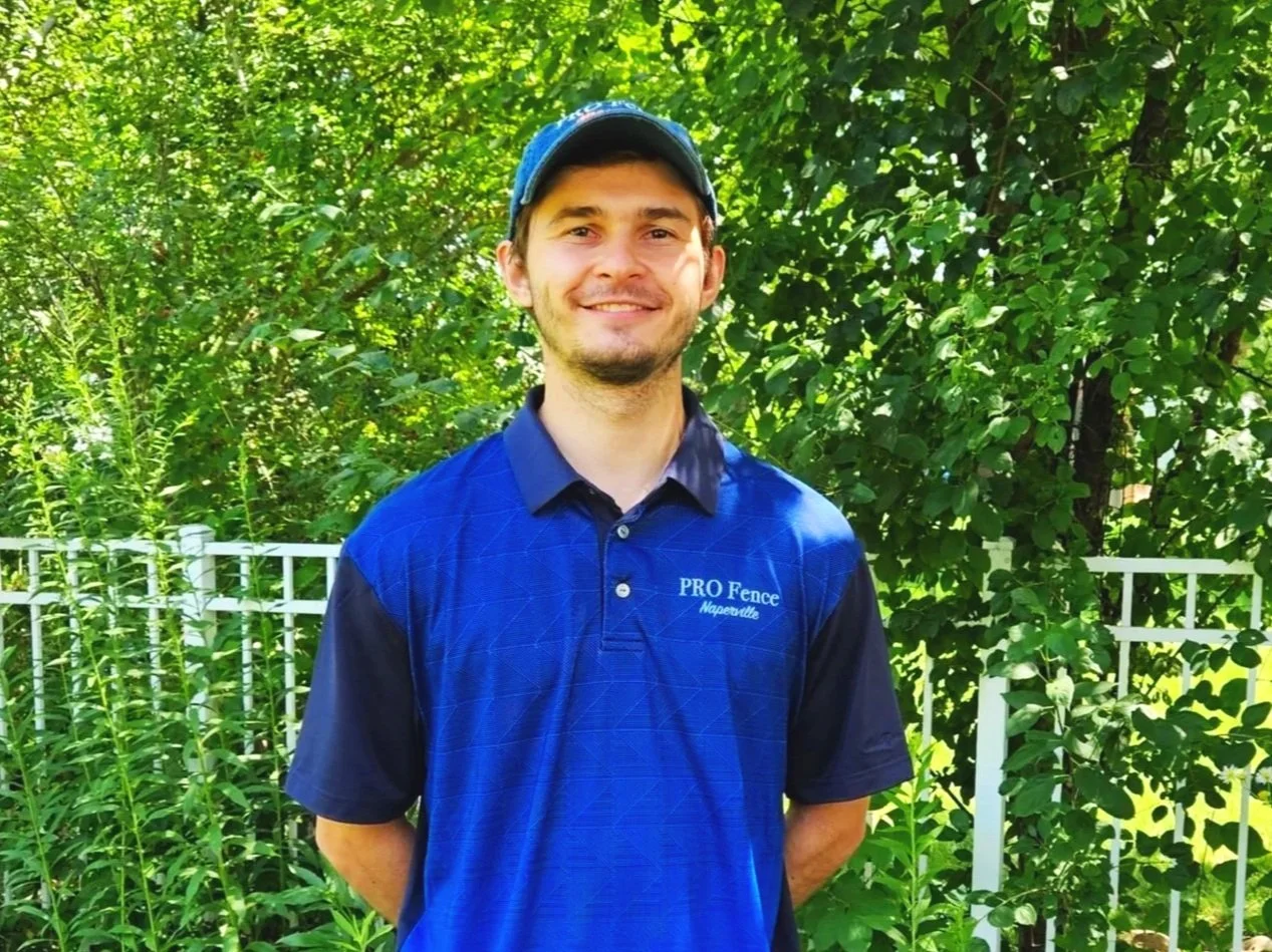 Smiling man wearing a blue cap and a blue Pro Fence uniform shirt standing in front of green foliage and a white metal fence.