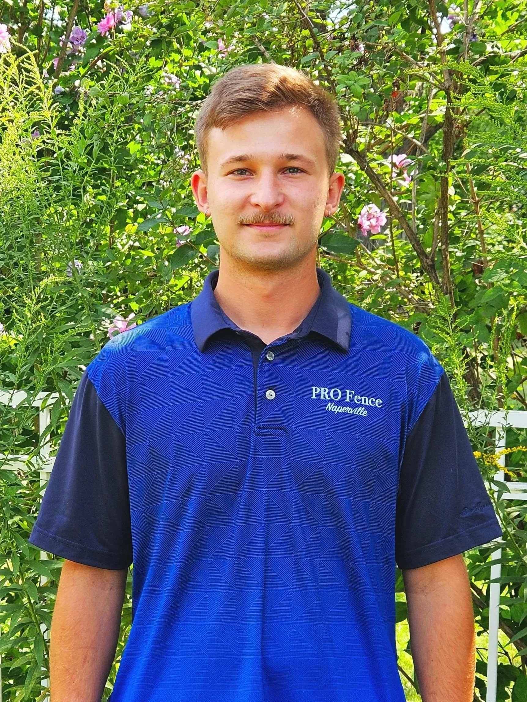 A young man in a polo shirt with 'PRO Fence Naperville' embroidered on it, stands outdoors in front of greenery and flowering plants, aluminum fence, and smiling