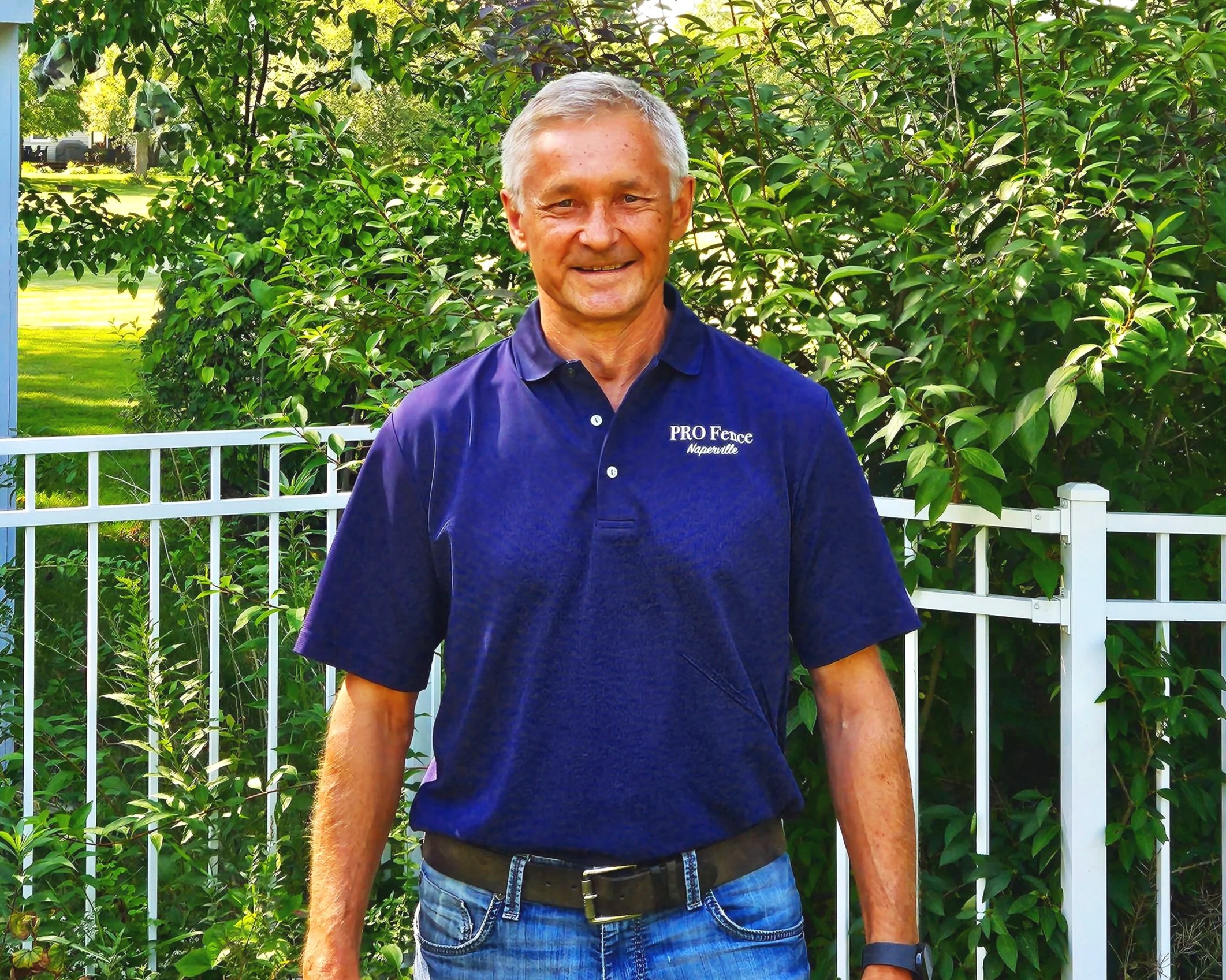 A smiling man with gray hair wearing a navy blue polo shirt and jeans stands outdoors in front of a white fence and green leafy trees. The polo shirt has white text that reads 'PRO Fence Naperville'.