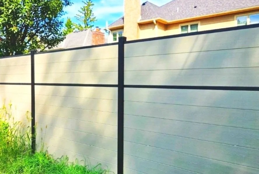 A modern composite privacy fence with black vertical posts and horizontal rails, adjacent to a grassy area with tall weeds.