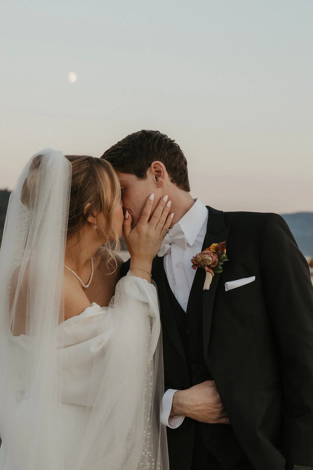 A bride and groom share a tender moment, touching foreheads and holding each other's faces, outdoors during sunset with a visible moon in the sky.