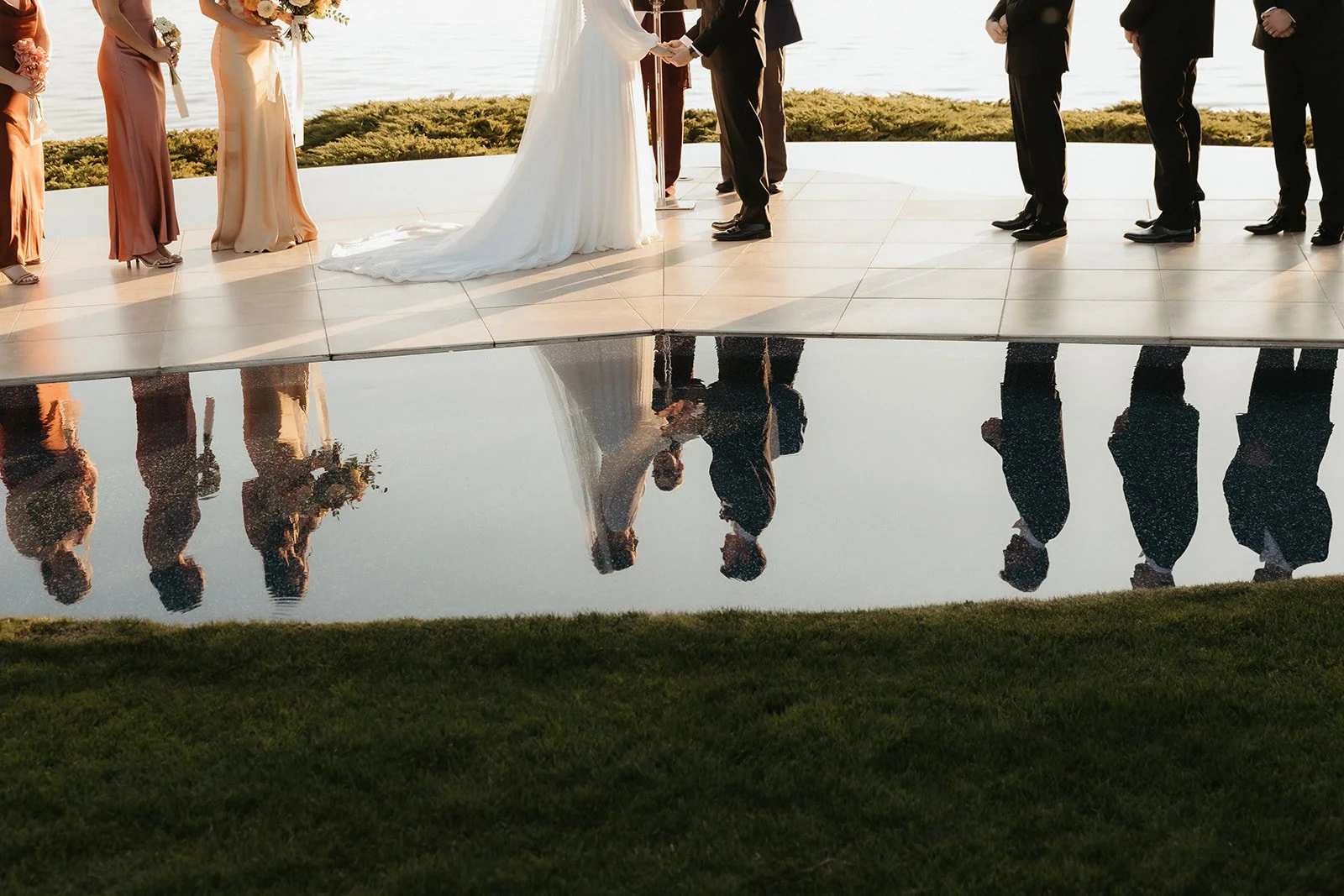 A wedding ceremony outdoors with a bride and groom exchanging vows, surrounded by bridesmaids and groomsmen, reflected in a water feature.