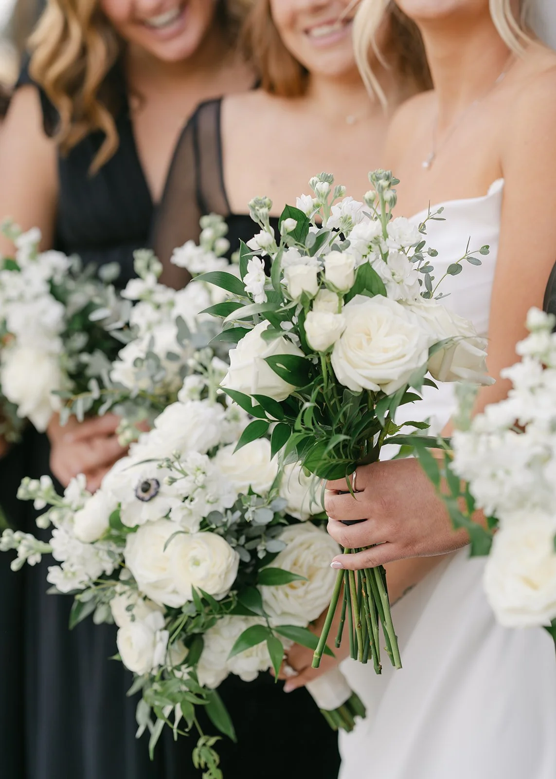 Close-up of a woman in a wedding dress holding a white floral bouquet at a wedding, with other women in black dresses and wedding attire in the background.