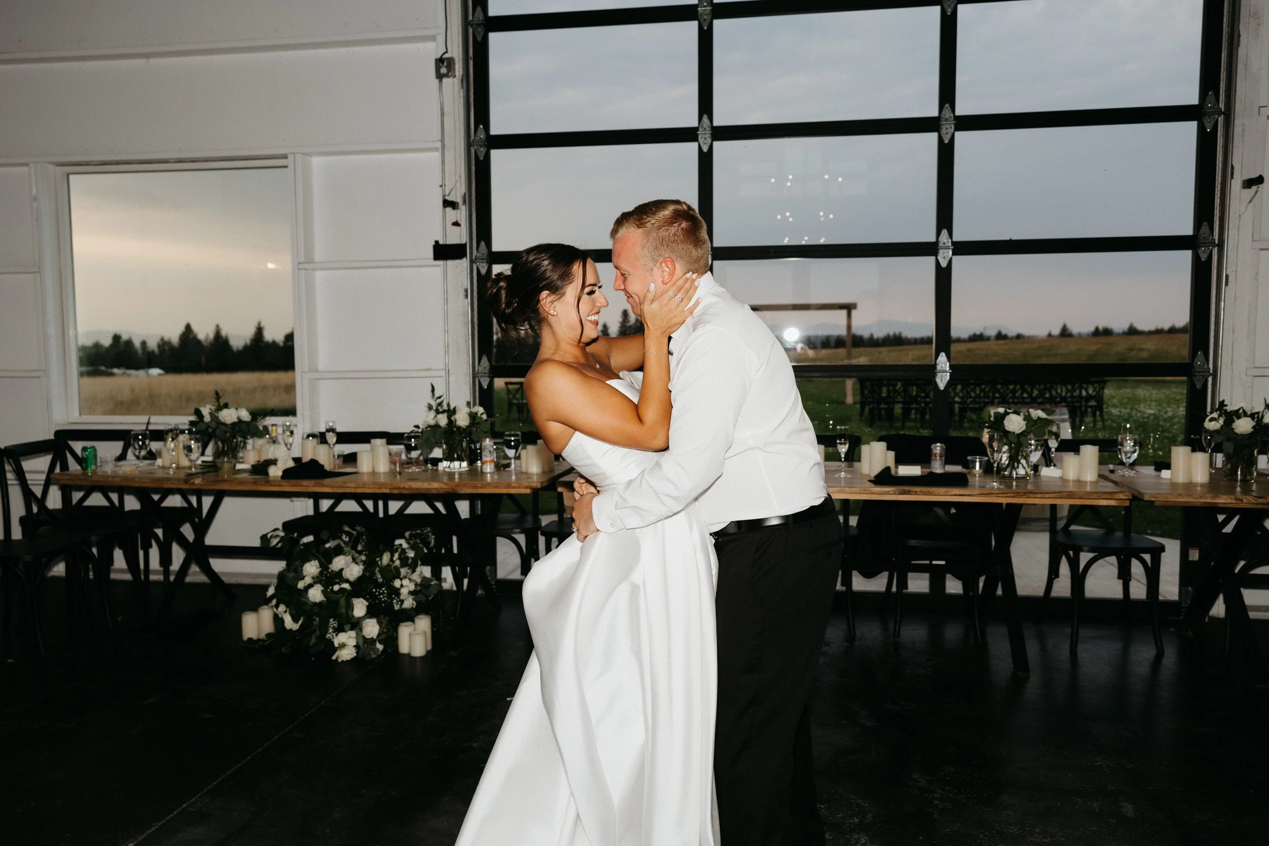 A bride and groom are dancing closely, smiling, during their wedding reception in a barn-like venue with large windows, decorated with white flowers and candles.