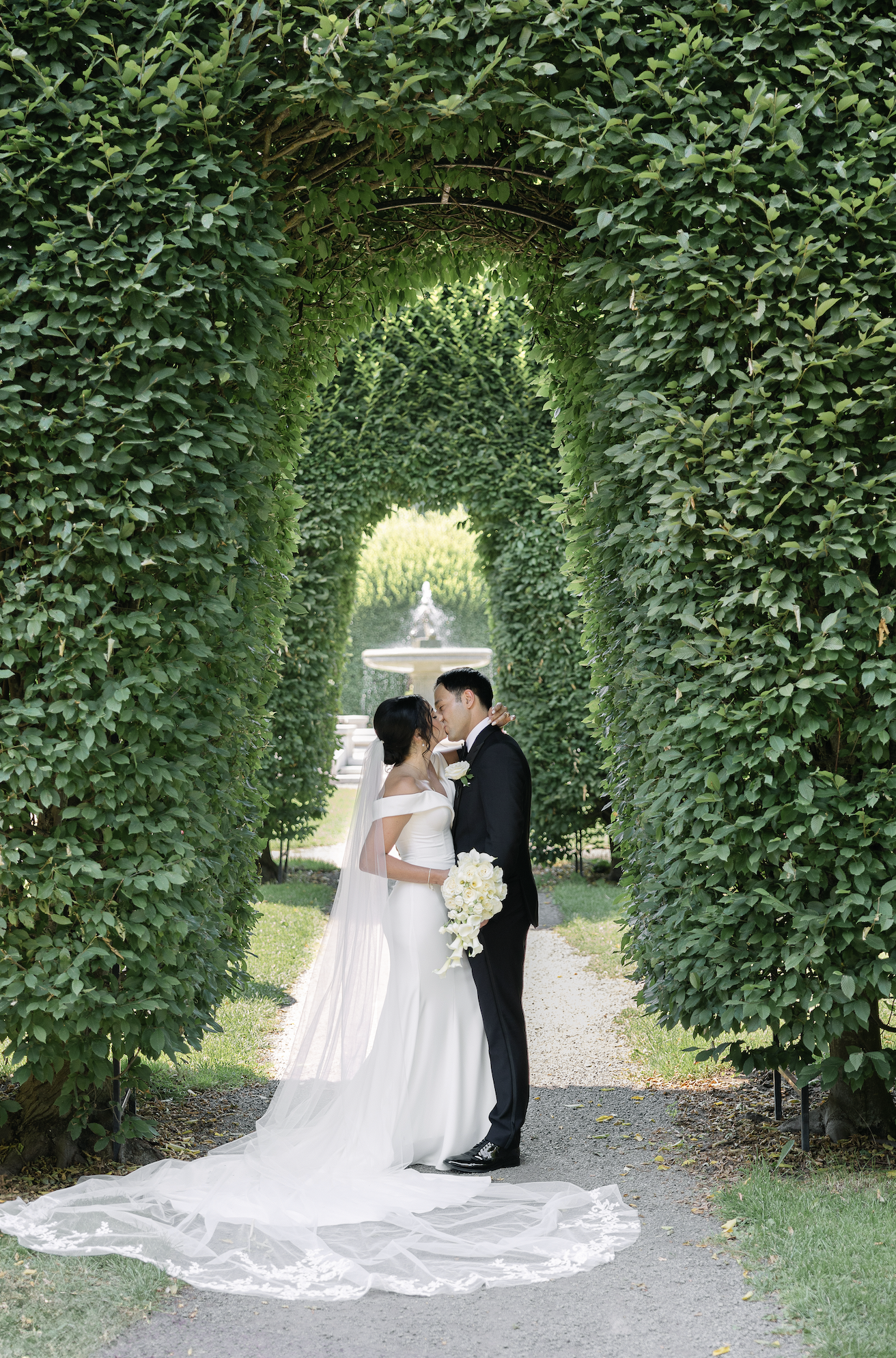 A bride and groom sharing a kiss under a green leafy arch in a garden, with a fountain visible in the background.