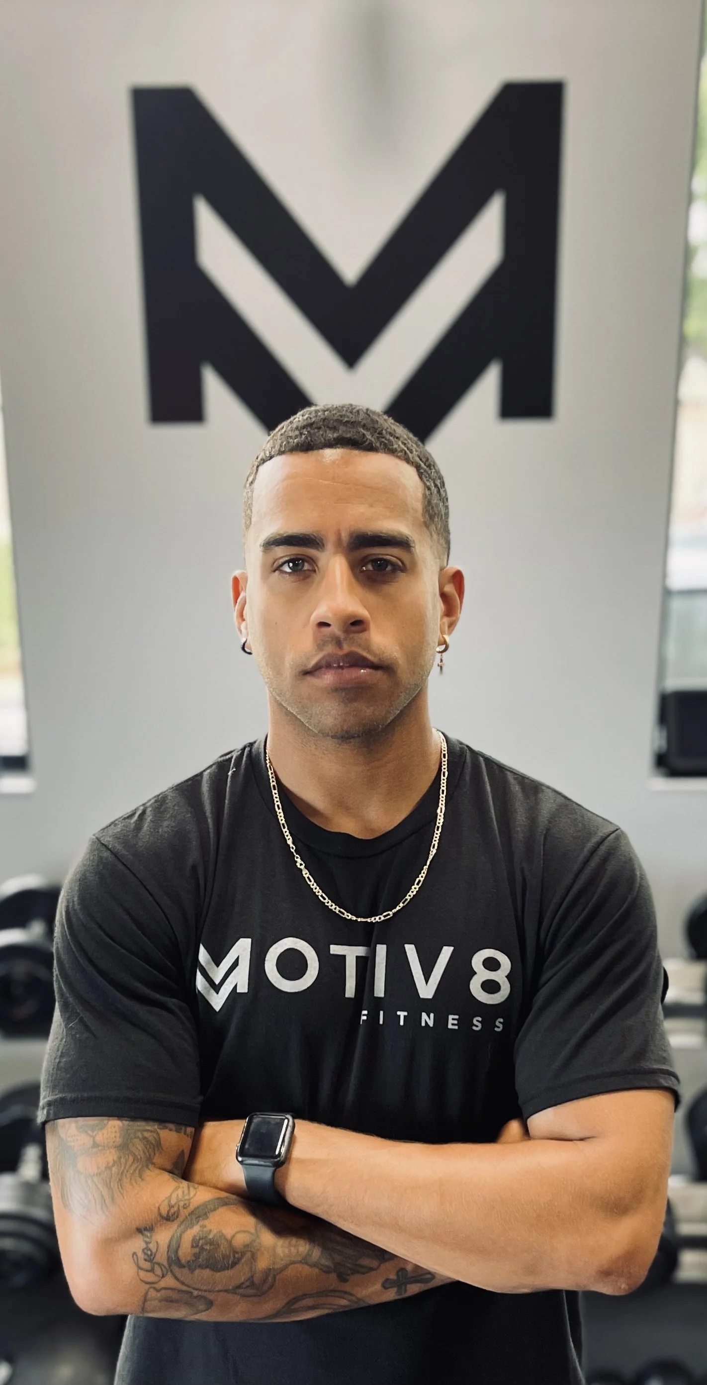 A young man with short dark hair and earrings, wearing a black Motiv Fitness T-shirt, a gold chain necklace, and a smartwatch, stands with crossed arms in a gym, with large black and white Motiv Fitness logo on the wall behind him.