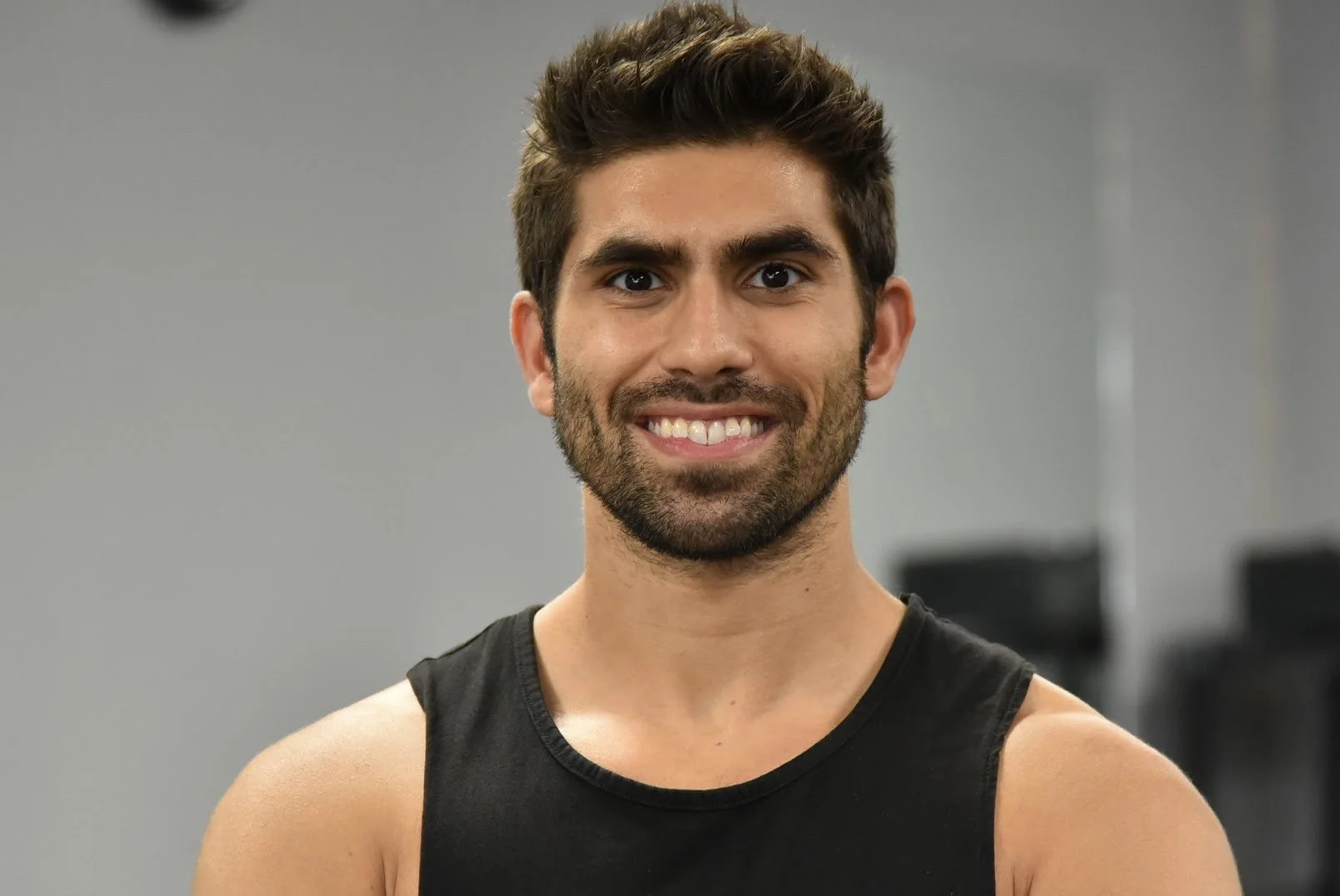 Young man with dark hair, beard, wearing a sleeveless black shirt, smiling indoors.