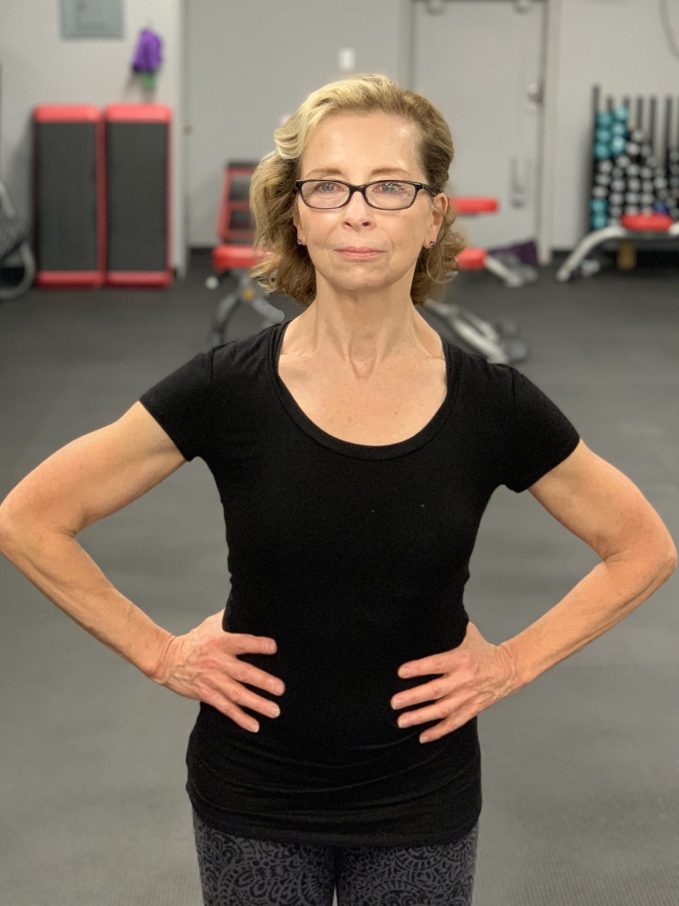 A middle-aged woman with blonde hair, glasses, and light skin standing in a gym with her hands on her hips, wearing a black t-shirt and patterned leggings.