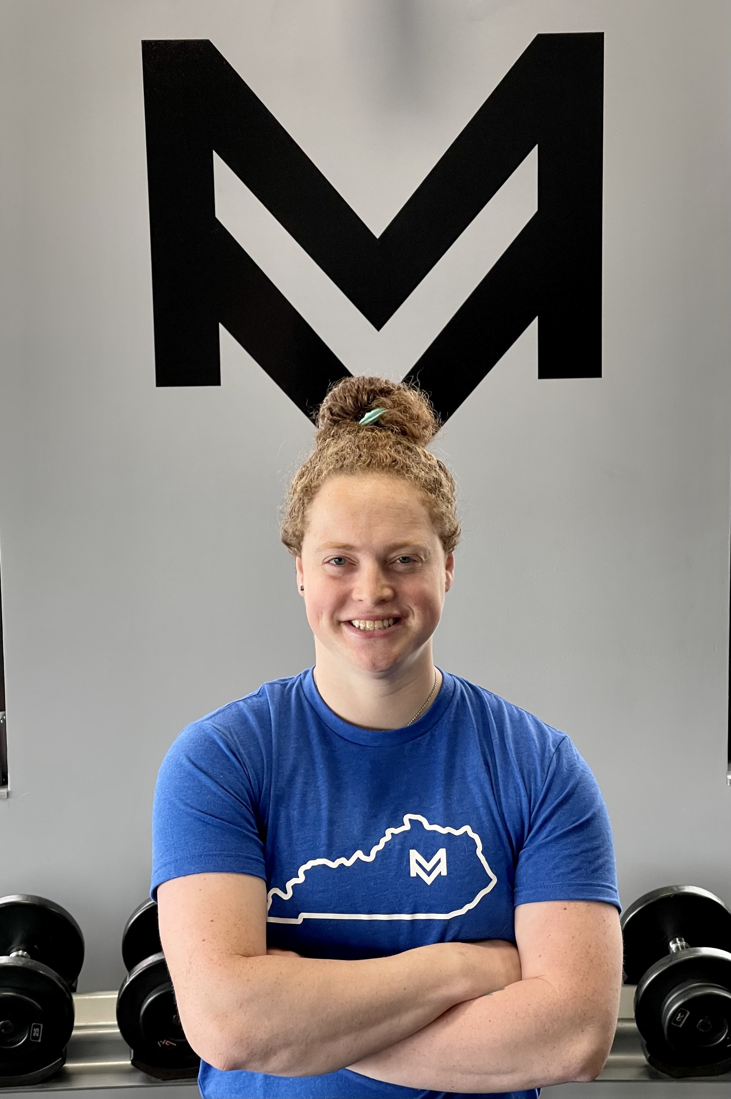 A woman with curly red hair tied in a bun, smiling and standing with arms crossed, in front of a gym wall with large black logo. She is wearing a blue T-shirt with a white outline of Kentucky and an M inside it, and there are dumbbells on a rack behind her.