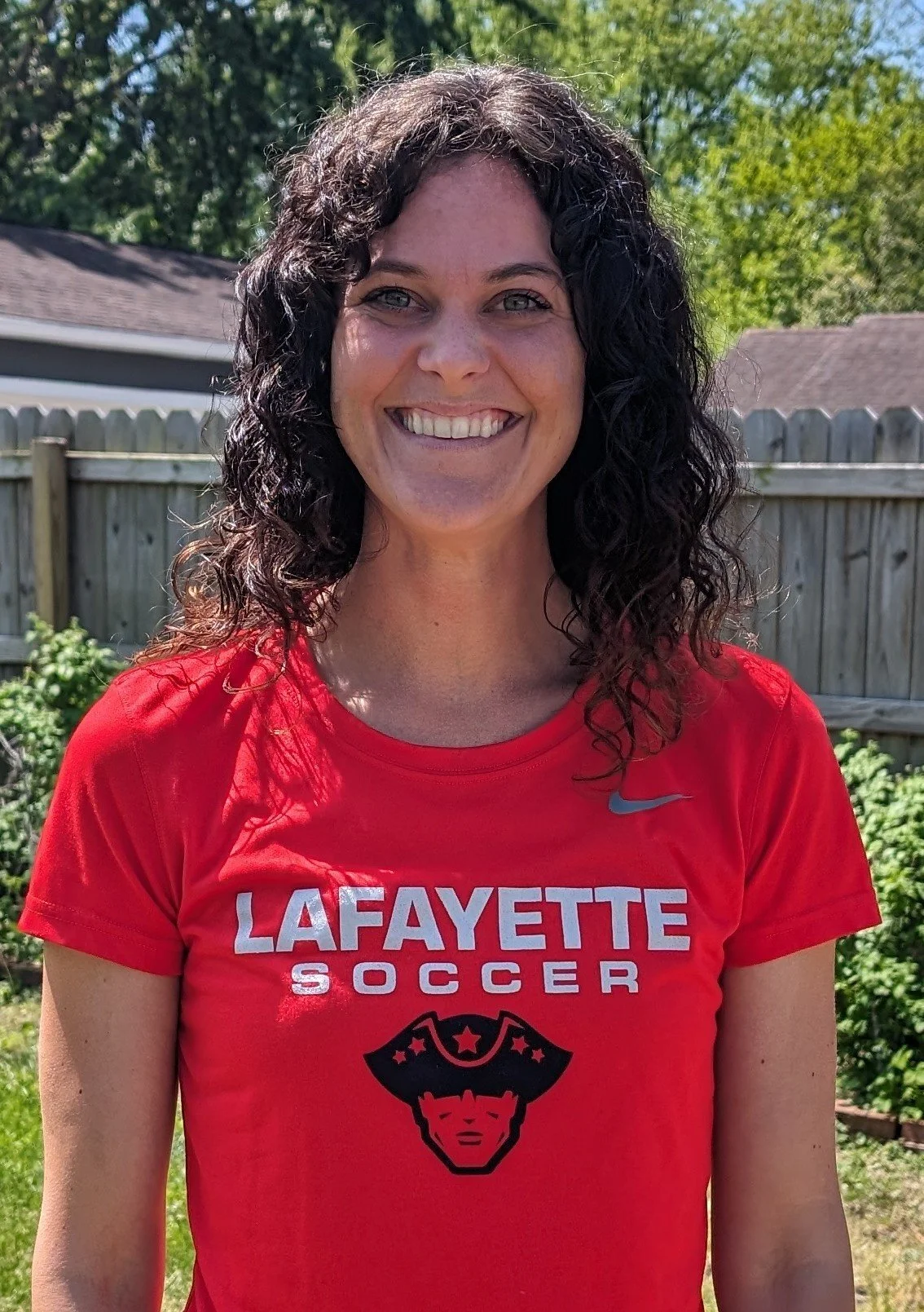 A woman with curly dark hair smiling outdoors in front of a wooden fence and green trees, wearing a red Lafayette Soccer T-shirt.