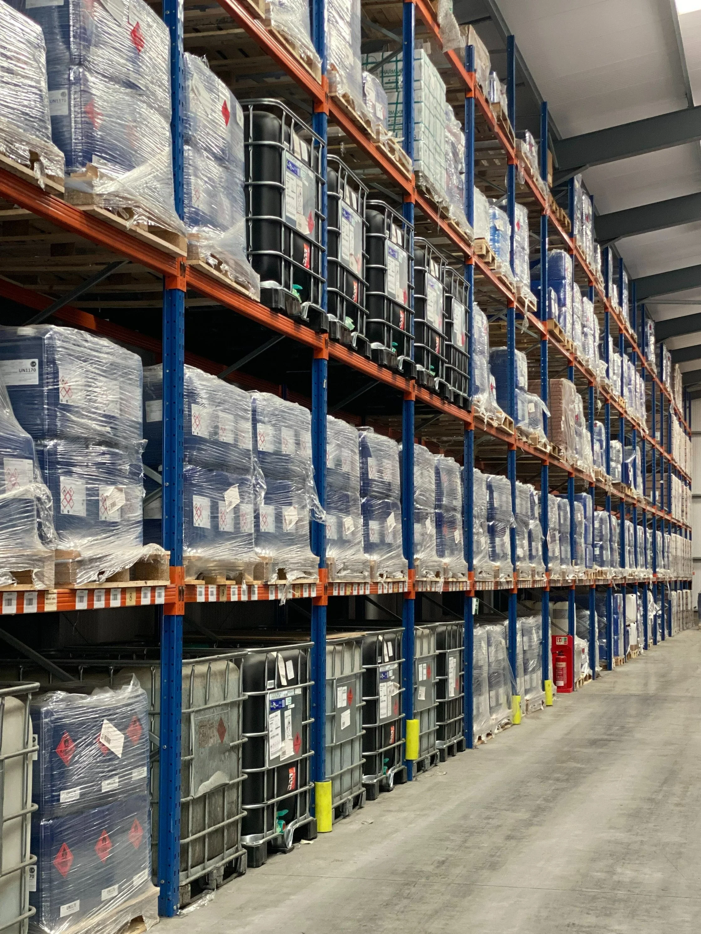 Warehouse aisle with shelves stacked with large containers and pallets wrapped in plastic.