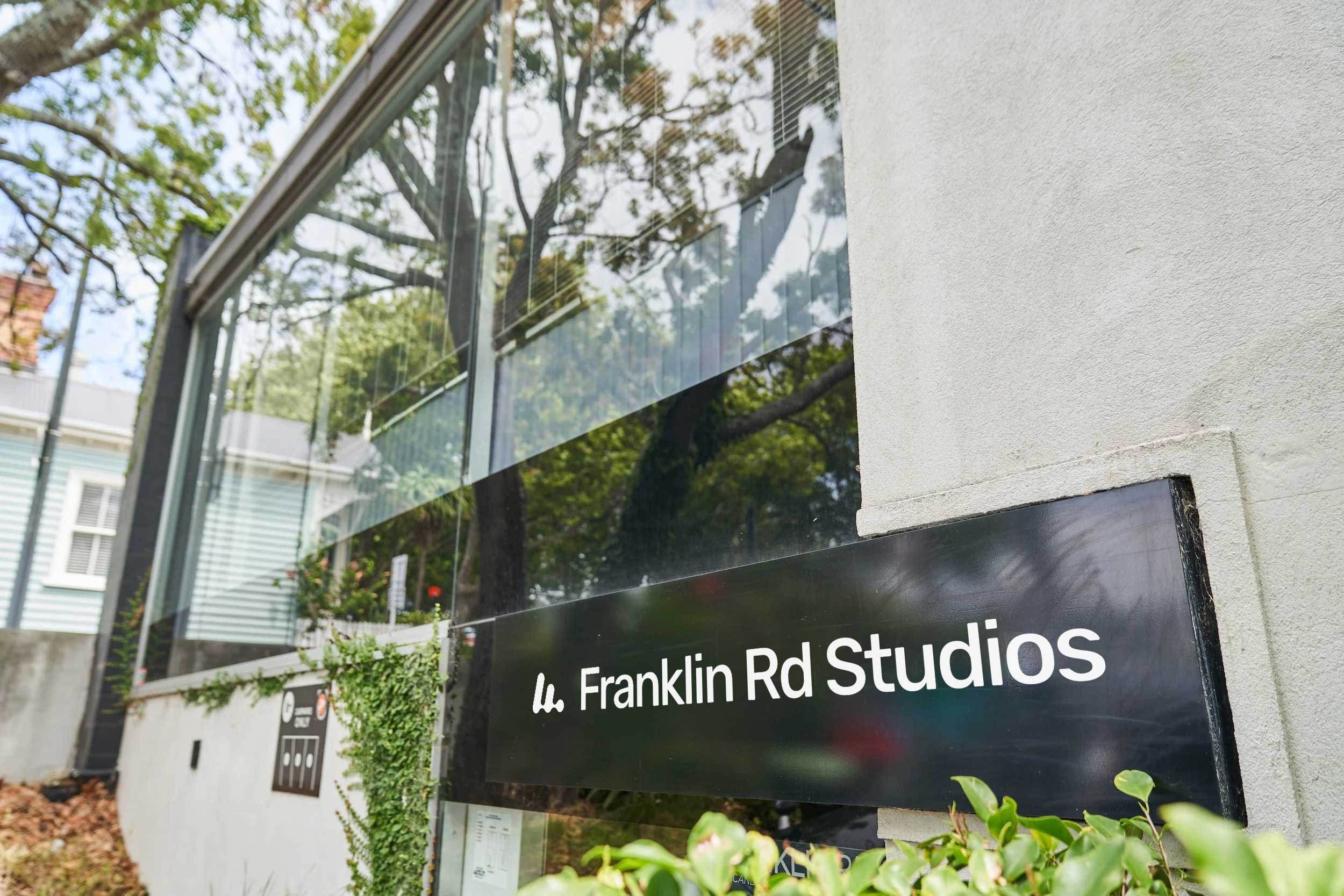 A contemporary studio exterior featuring a sleek black signage panel mounted against a textured white façade. The sign displays the Franklin Rd Studios logo in crisp white. Framed by lush greenery and reflected tree canopies in the expansive glass wi