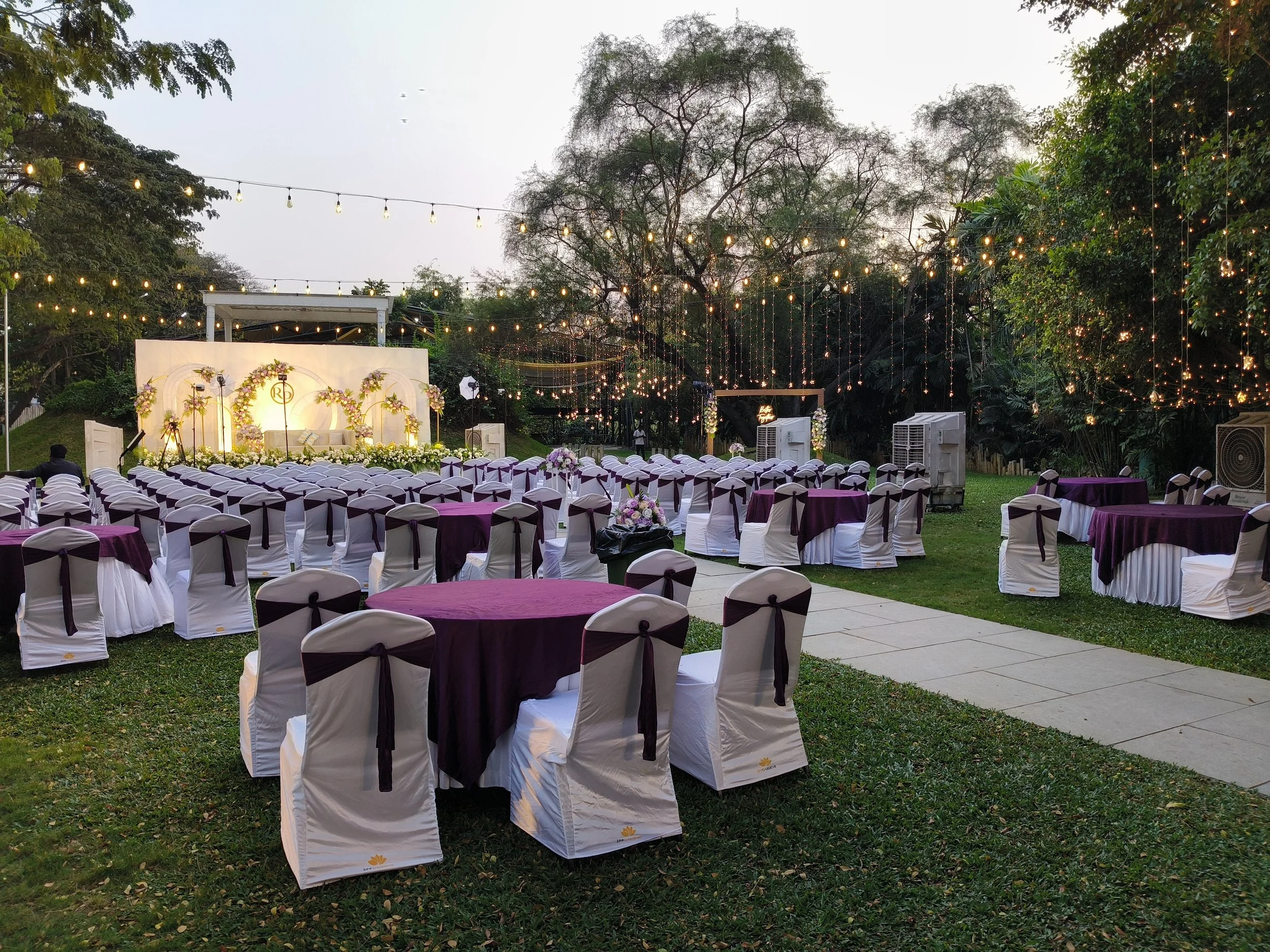 Outdoor wedding setup at dusk with round tables purple linens fairy lights strung across trees and a floral lit stage