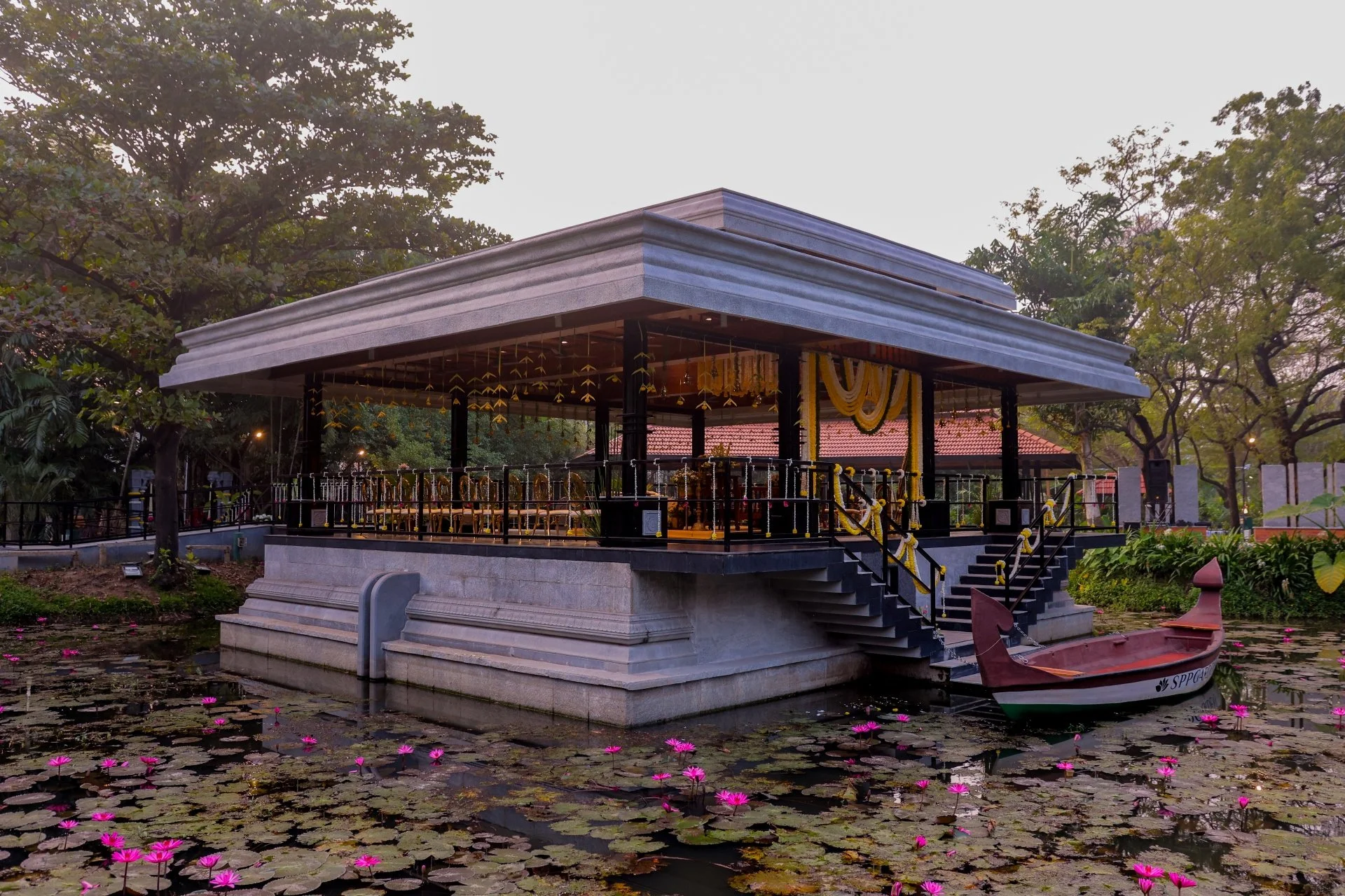 Stone pavilion with sloped roof surrounded by lily pond with pink water lilies and a wooden boat docked at the steps