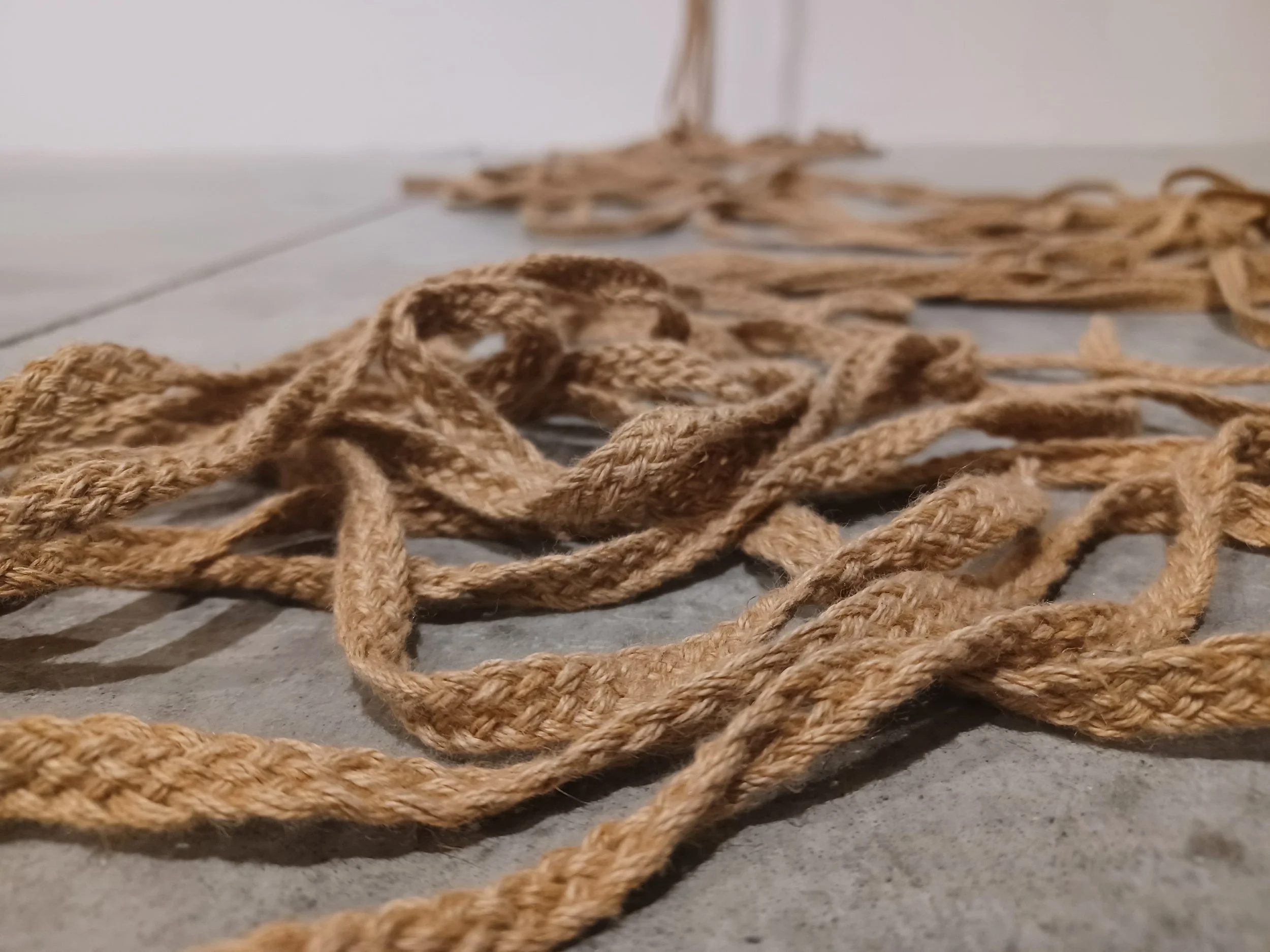 Close-up of braided camel-coloured rope trailing from a white wall onto the grey floor, pooling towards the viewer.