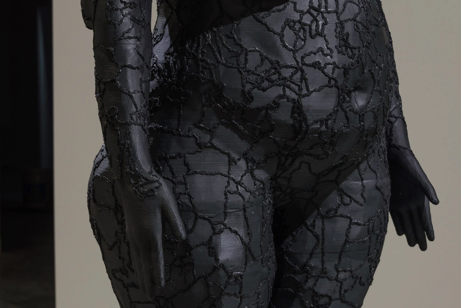 Close-up of a black 3D-printed sculpture of the artist, focusing on the torso, arms, and hands. The sculpture features intricate embroidery on the surface, giving it a textured look.