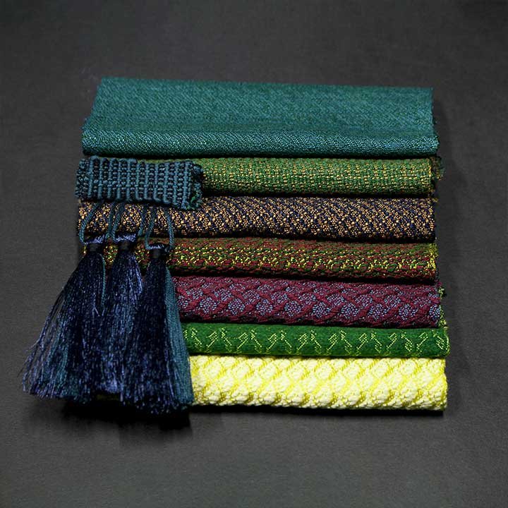 A neatly stacked pile of seven colorful, patterned woven fabric pieces with passementerie tassels on one end, laid on a black surface.