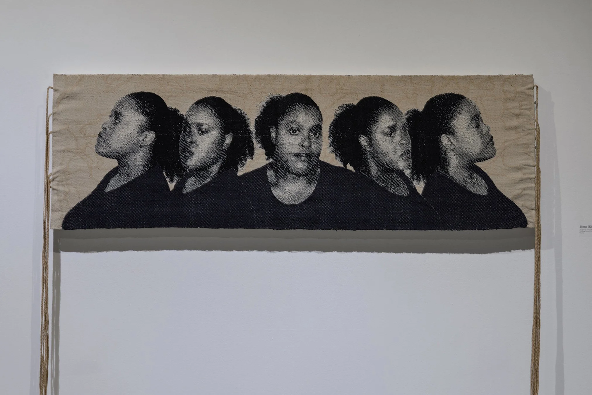 Woven horizontal tapestry installation featuring five black and white portrait photographs of the artist in profile, with ropes trailing from the sides, against a plain white wall in a gallery.