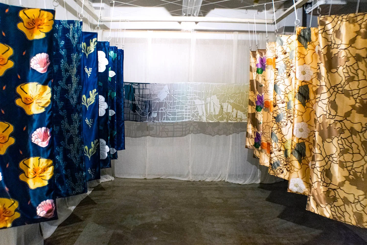 Printed drapes hanging in an indoor space, with a white mesh netting covering the wall behind. Flanked by the drapes and in front of the wall is a dyed burnout map.