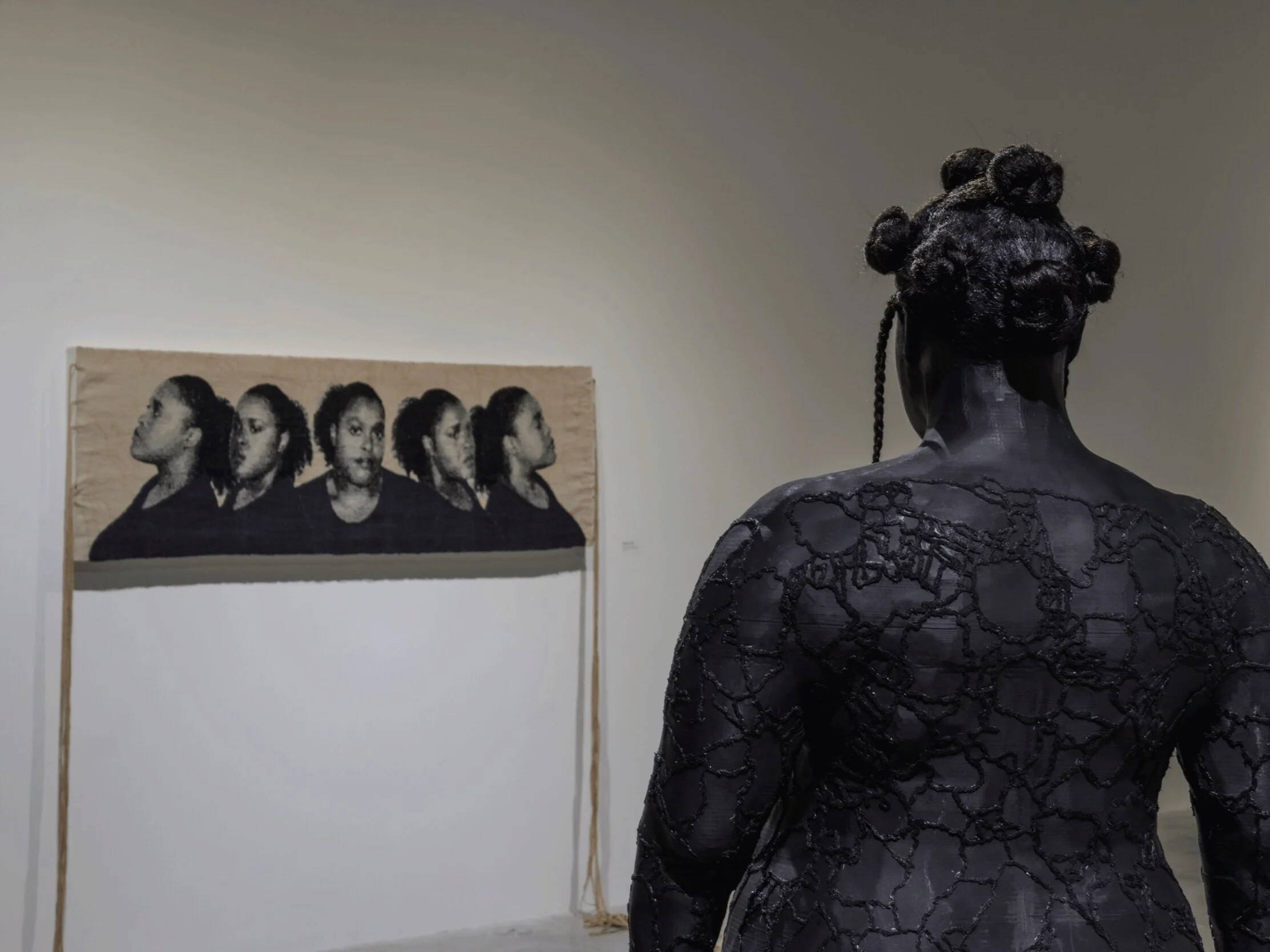 A 3D-printed sculpture with hair styled in bantu knots faces away from the viewer, towards a tapestry on the wall. The sculpture features intricate embroidery on the surface.