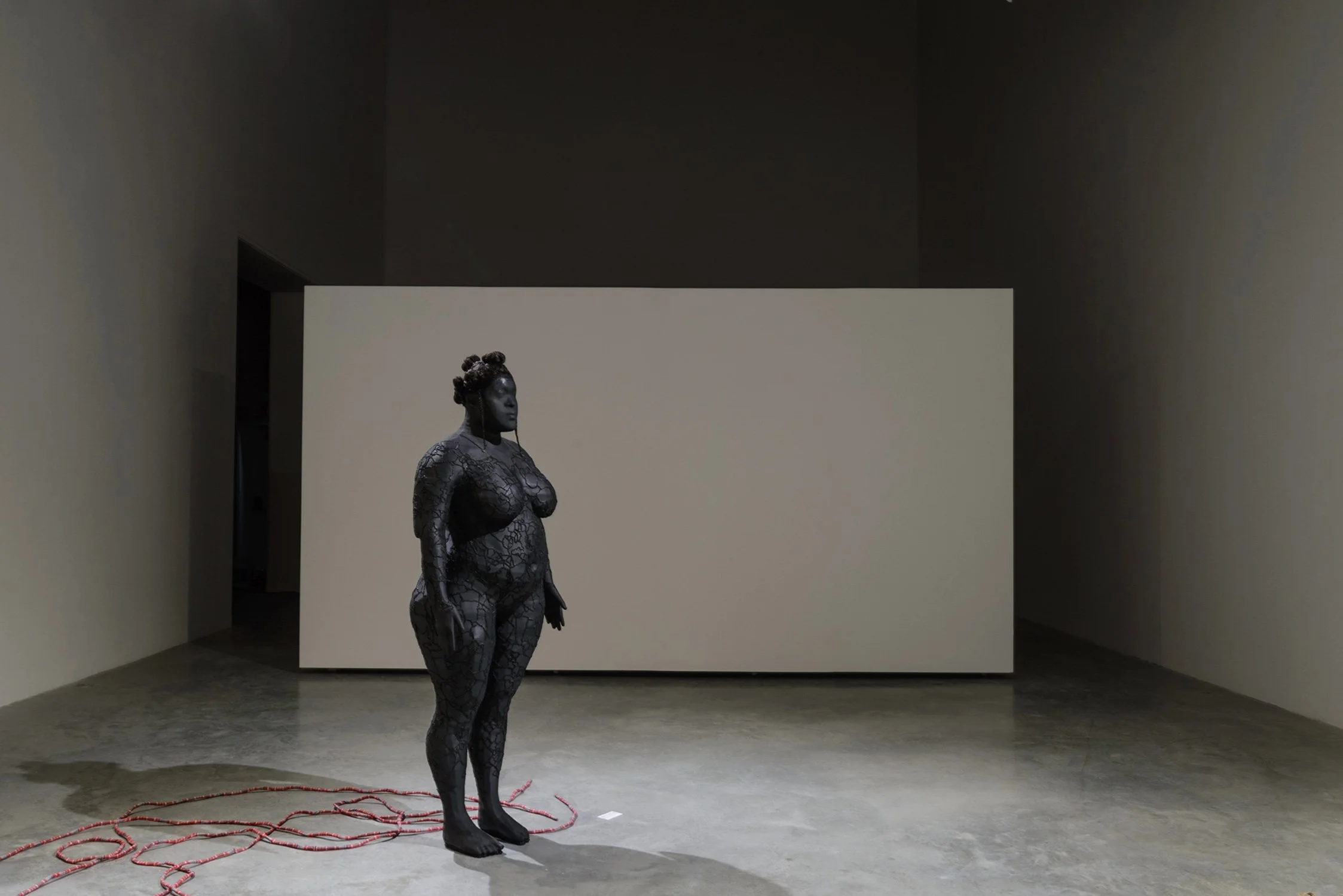 A life-size black 3D-printed statue of Shiemara with curly hair, standing on a smooth concrete floor inside a gallery. The body is covered in black embroidery. Behind the statue, there is a large white wall in a dimly lit room with beige wall.