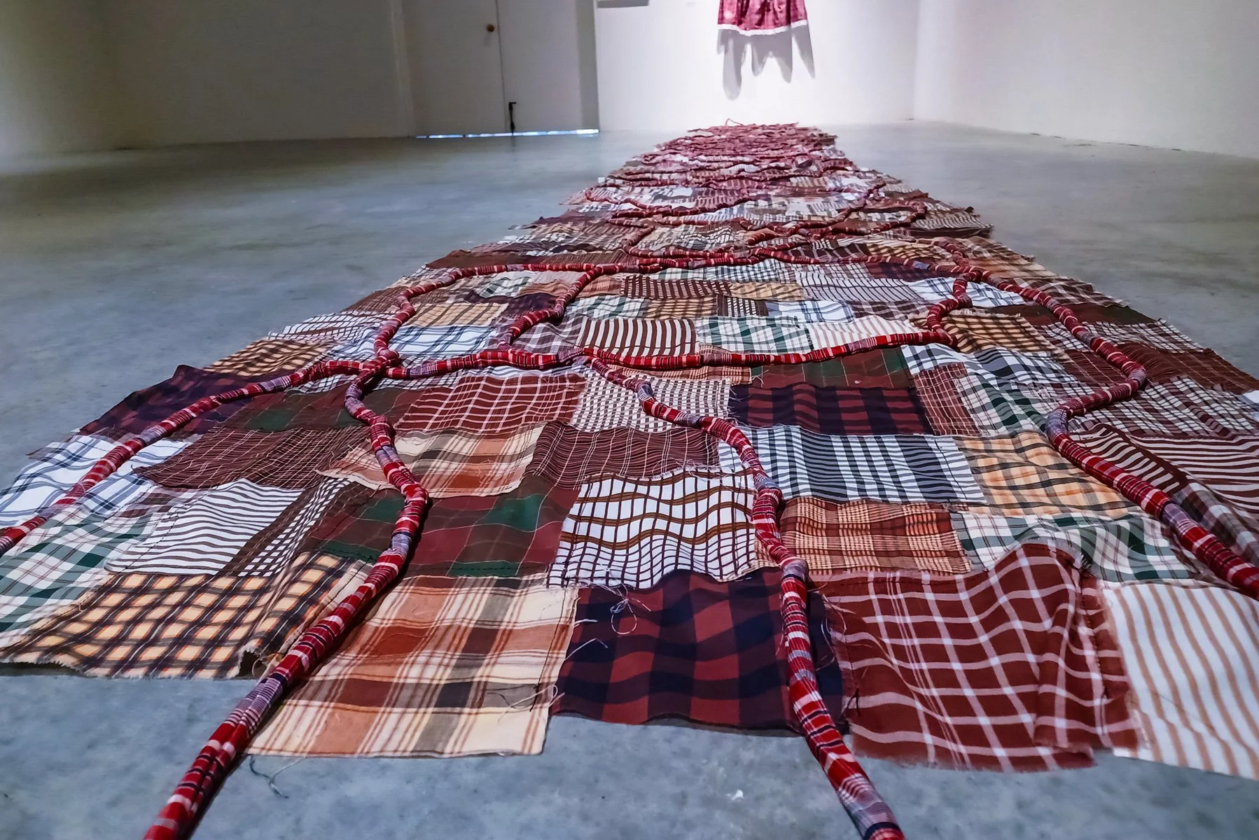 Installation featuring a long patchwork quilt of various plaid fabric squares arranged on a concrete floor, with red and pink striped borders, in an art gallery setting.