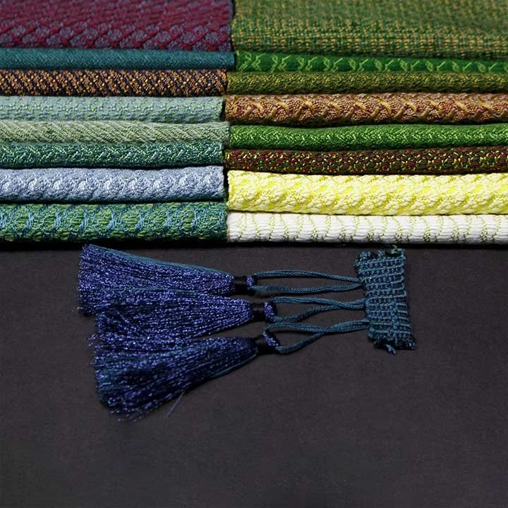 Stacked colorful woven fabric samples with a blue passementerie fringe on a black surface.