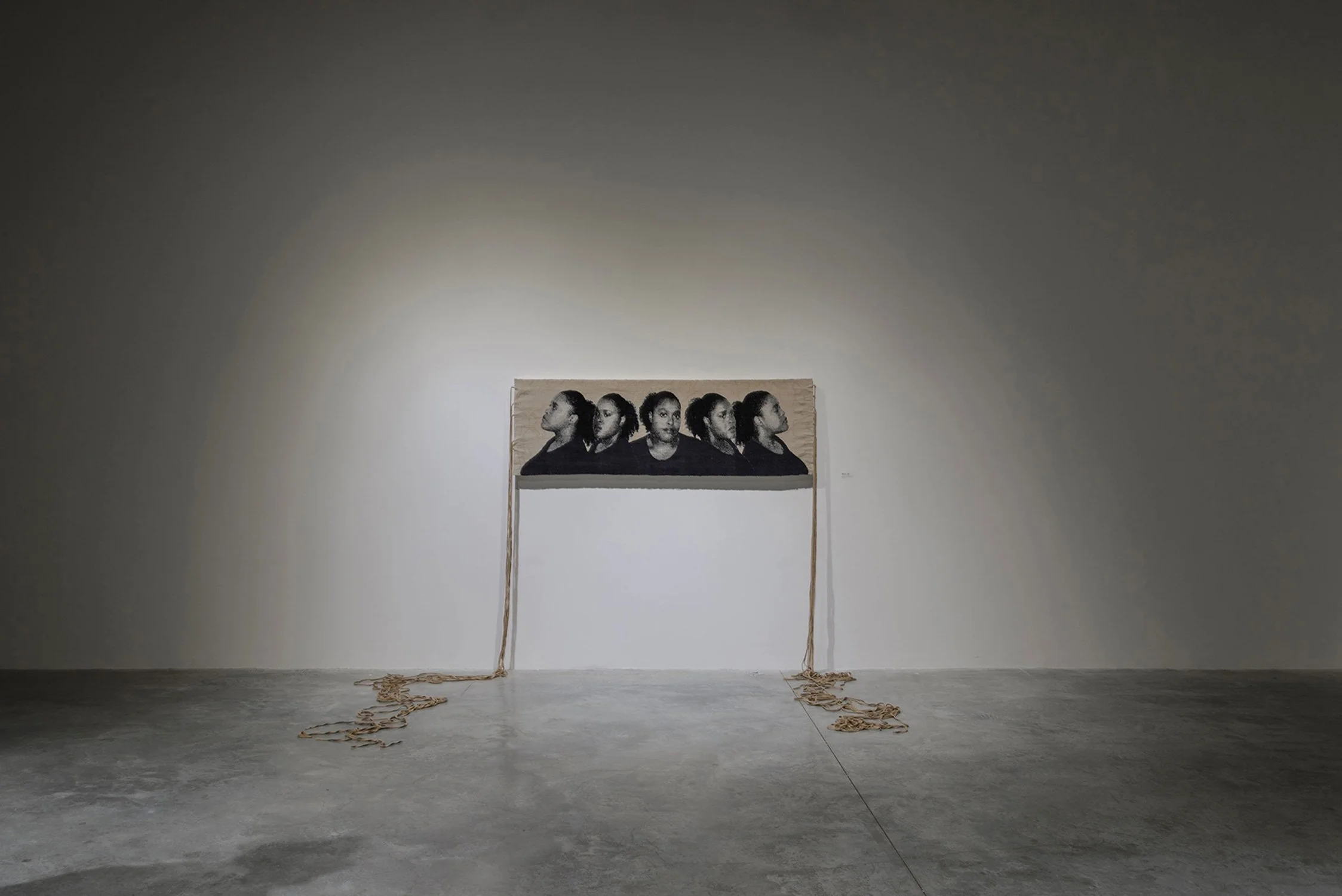 Woven horizontal tapestry installation featuring five black and white portrait photographs of the artist in profile, with ropes trailing from the sides, against a plain white wall in a gallery.