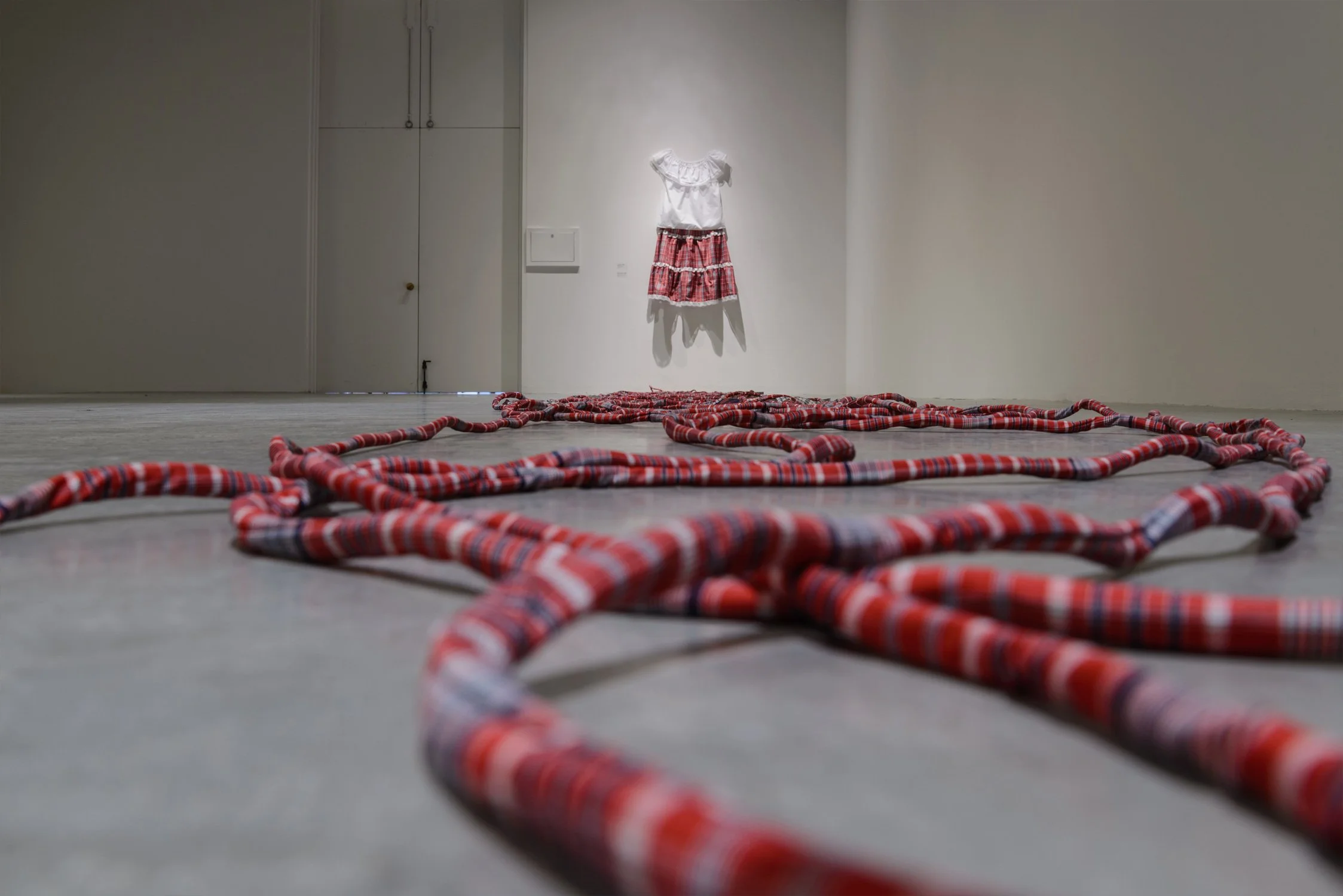 A gallery exhibit showing the Jamaican bandana outfit hanging on the wall - red plaid skirt and white top - and large red plaid ropes trailing toward the viewer on the floor in the foreground.