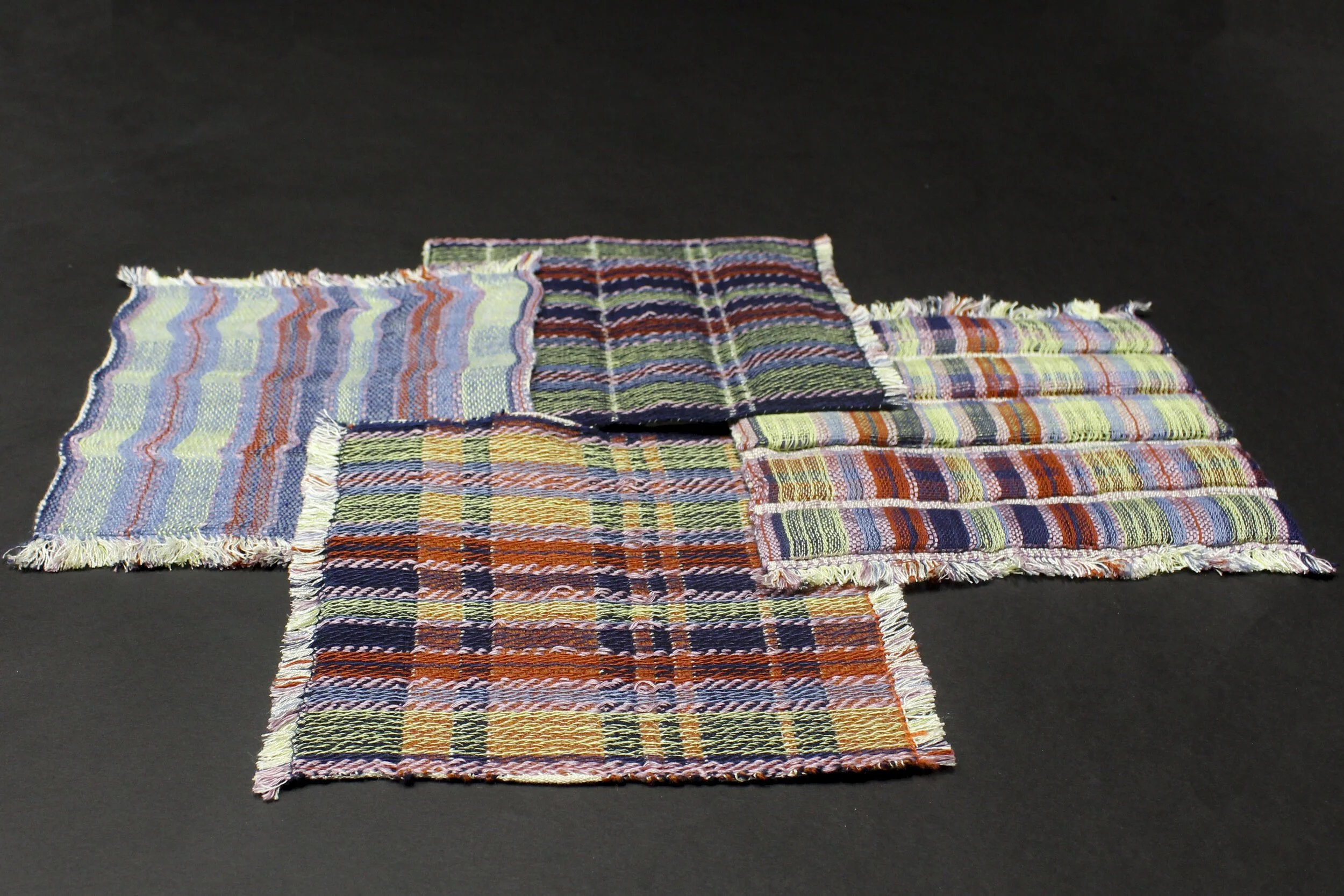 Six colorful woven doublecloth padded fabric square samples with various plaid and stripe patterns laid out on a black surface.