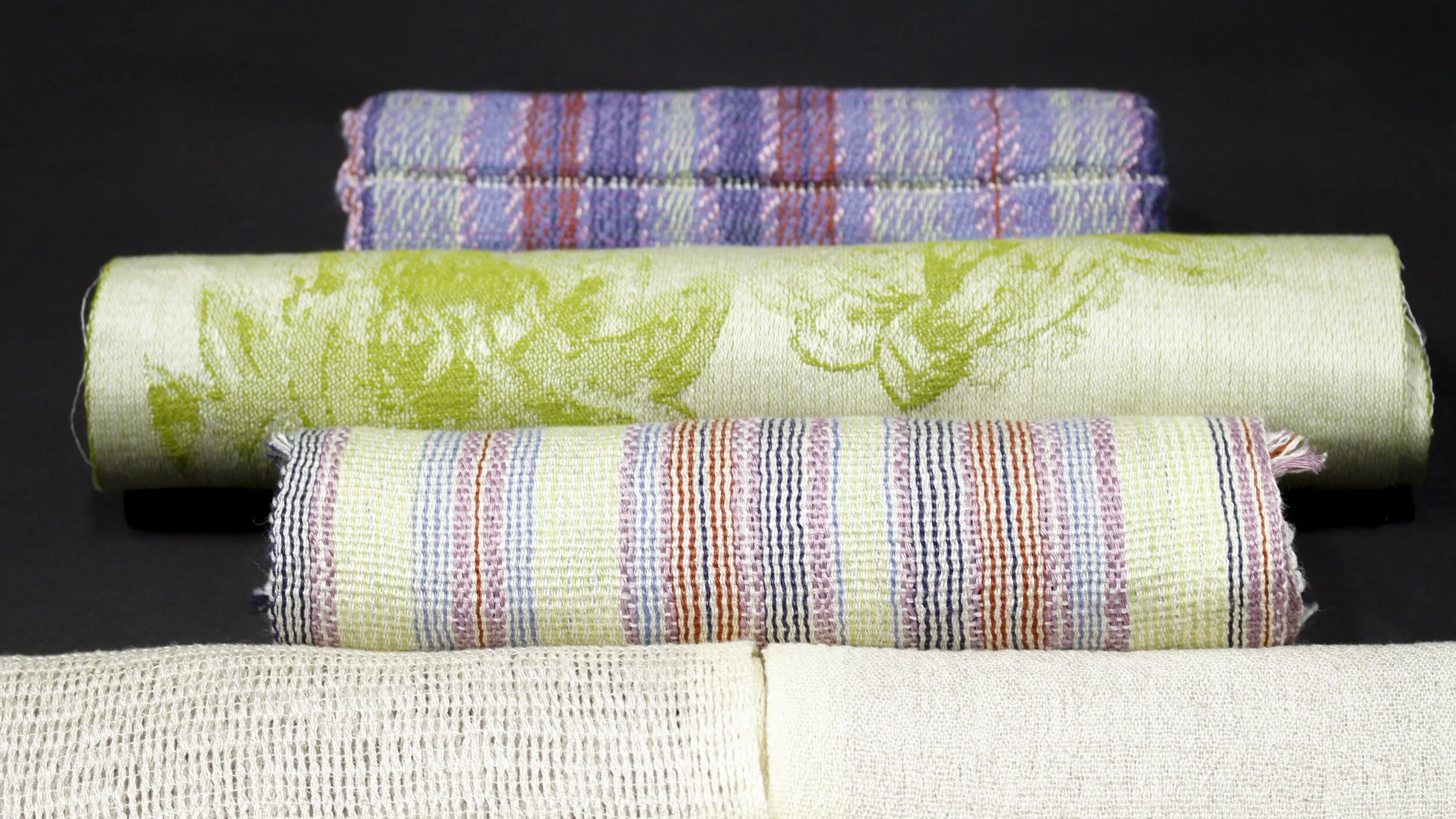 Rolled jacquard, leno lace and doublecloth samples of various patterns and colors, placed on a black surface.