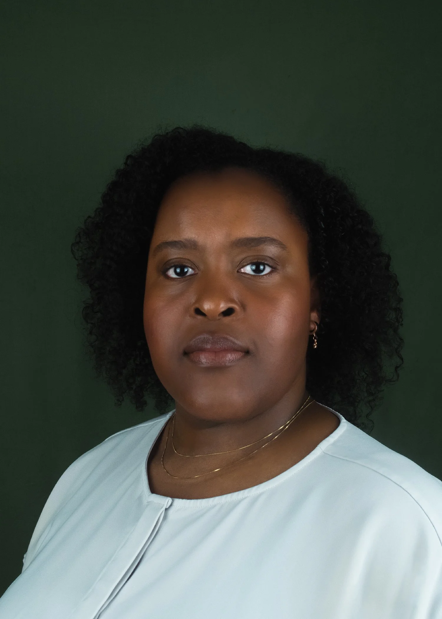 A profile photo of Shiemara with dark curly hair, wearing a light-colored top, gold jewelry, and earrings, looking directly at the camera against a dark green background.
