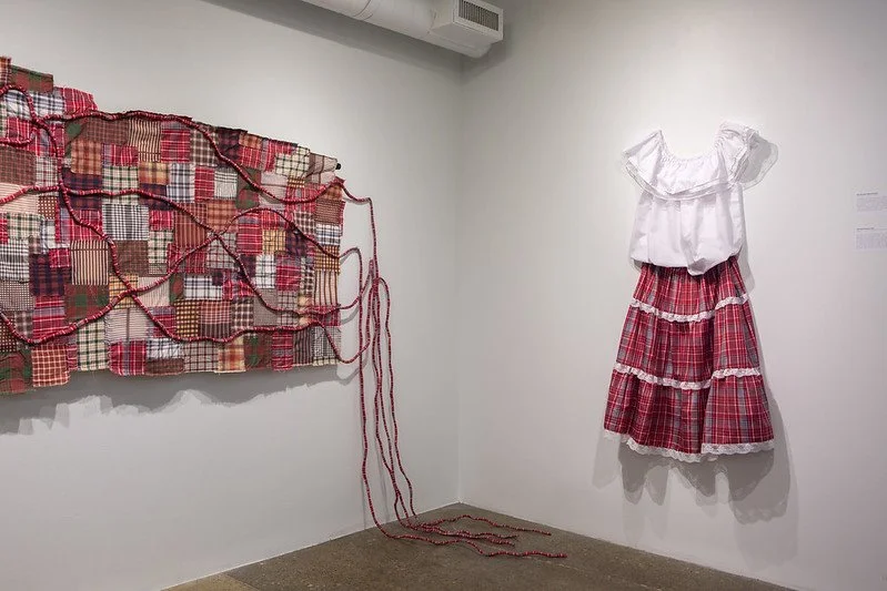 Mixed media art installation featuring a patchwork quilt mounted on the left wall with red rope trailing through it, and a white blouse paired with a layered red plaid skirt on the right wall displayed as art.