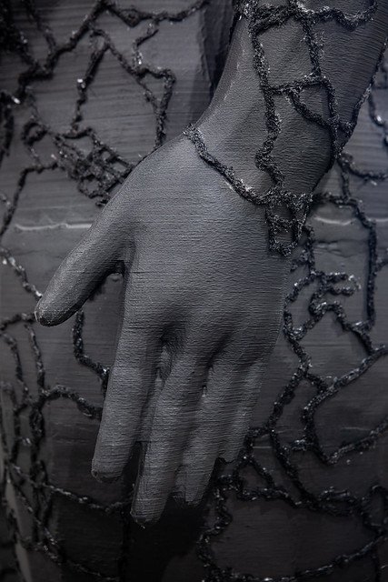 Close-up of a black 3D-printed sculpture of the artist, focusing on the left hand. The sculpture features intricate embroidery on the surface.