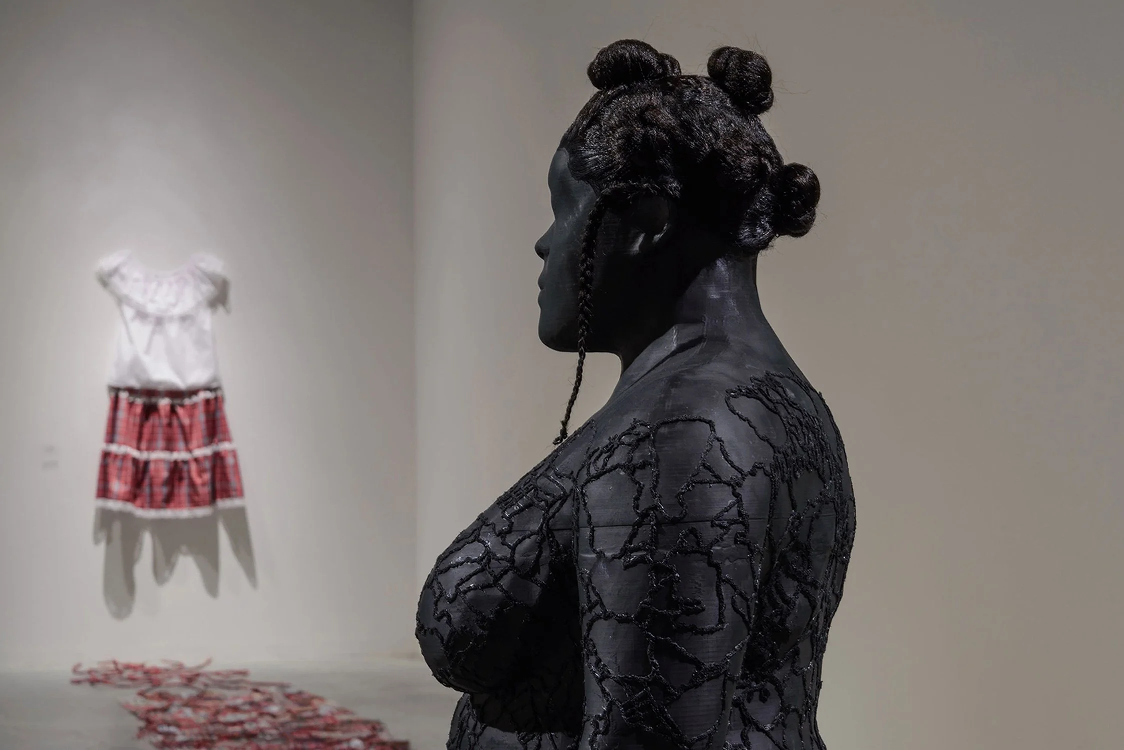 Black 3D-printed sculpture of a woman with hair styled in bantu knots, adorned with black embroidery, facing sideways in an art gallery. In the background, the Jamaican bandana outfit hangs against a wall, with a patchwork quilt on the floor.