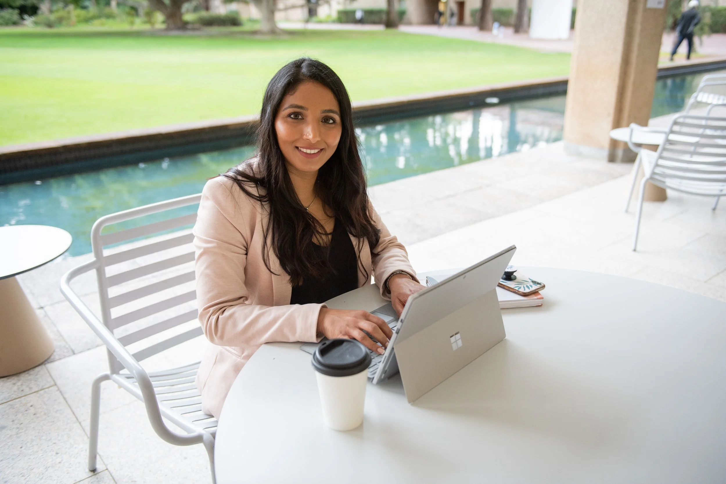 A woman with long dark hair sitting at a white outdoor table with a laptop, smiling, with a green park background.