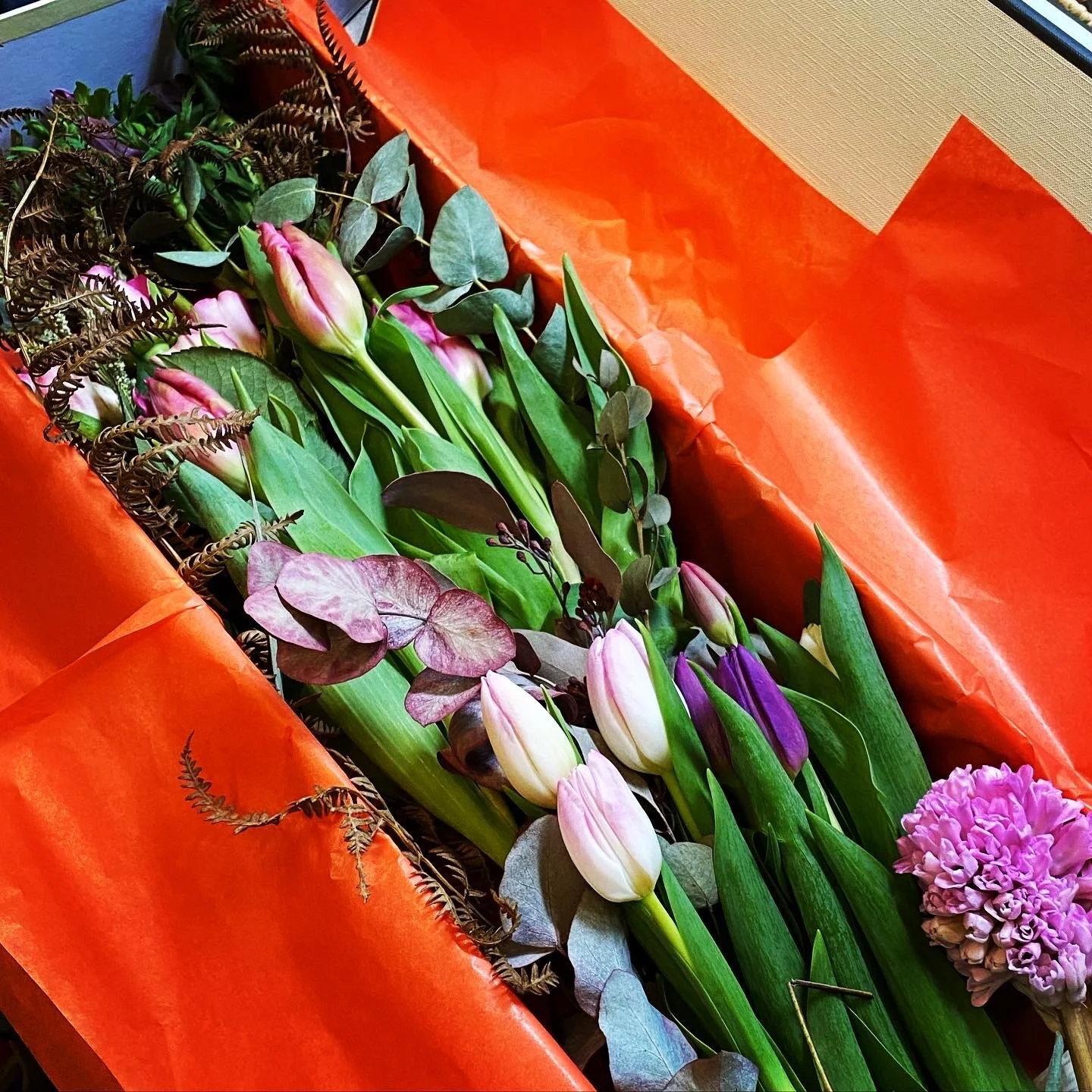 A bouquet of pink, purple, and white tulips with green leaves, nestled in orange wrapping paper with some dried brown foliage and purple filler flowers.