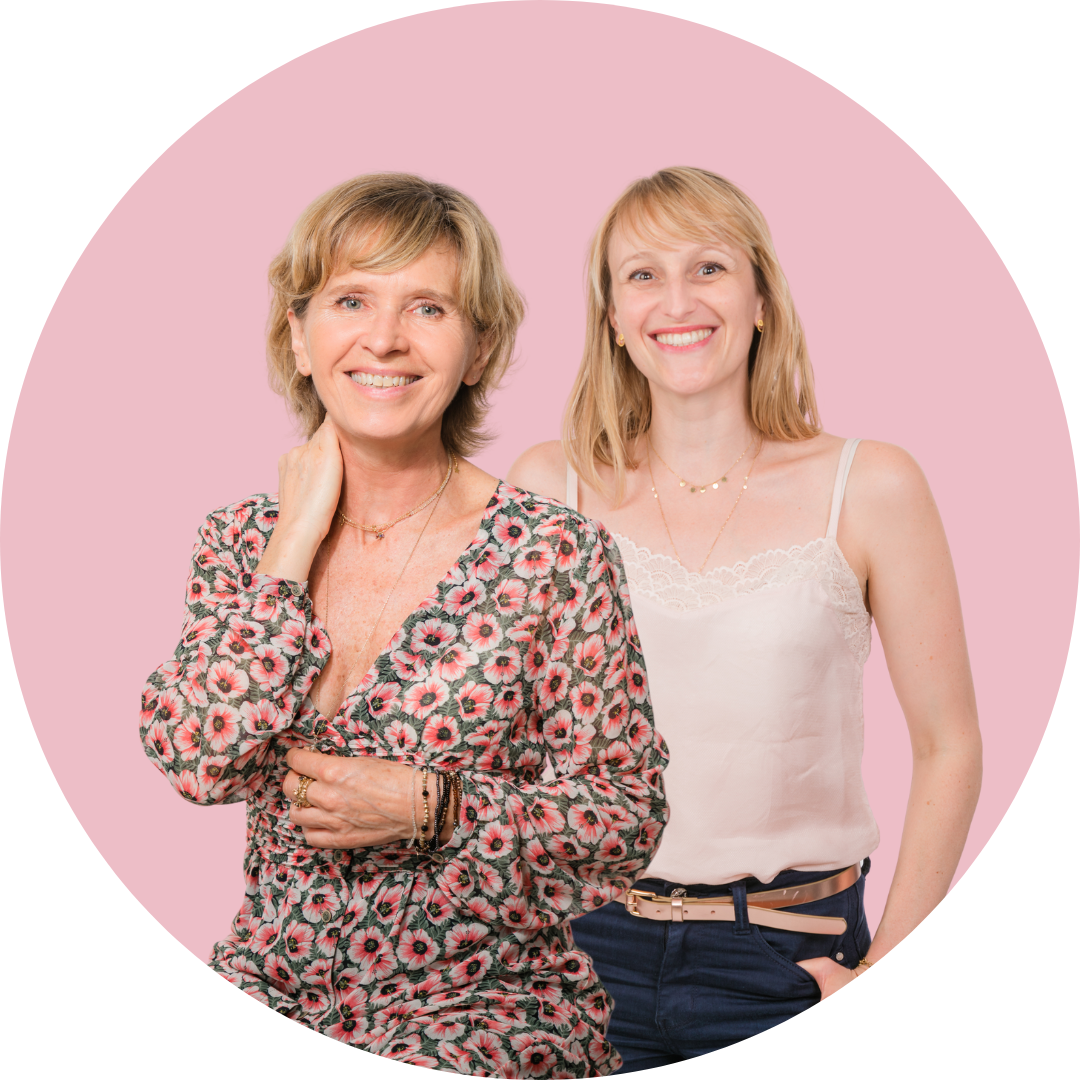 Deux femmes souriantes posant devant un fond rose, l'une portant une robe à motifs floraux et l'autre une camisole blanche.