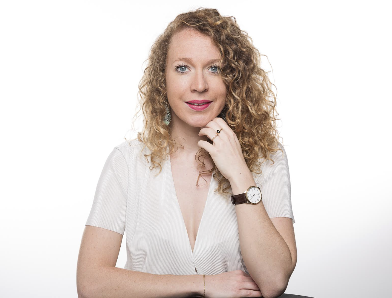 Portrait d'une femme aux cheveux bouclés, portant une montre et des bijoux, en blanc, souriante, sur fond blanc.