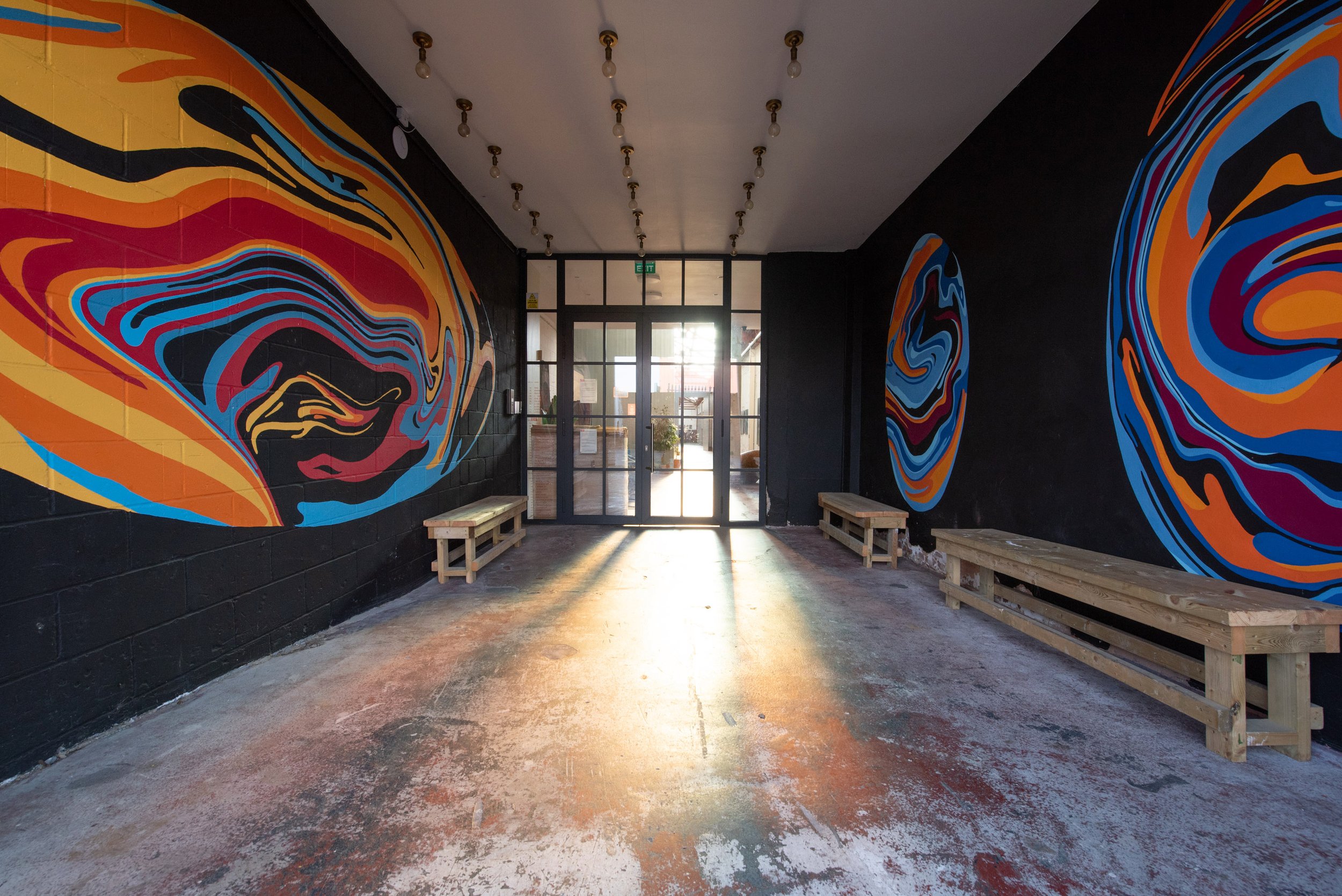 hallway Entrance to Excelsior Studios with black walls decorated with abstract, colourful swirl murals, wooden benches, concrete floor, and glass doors at the end letting in sunlight.