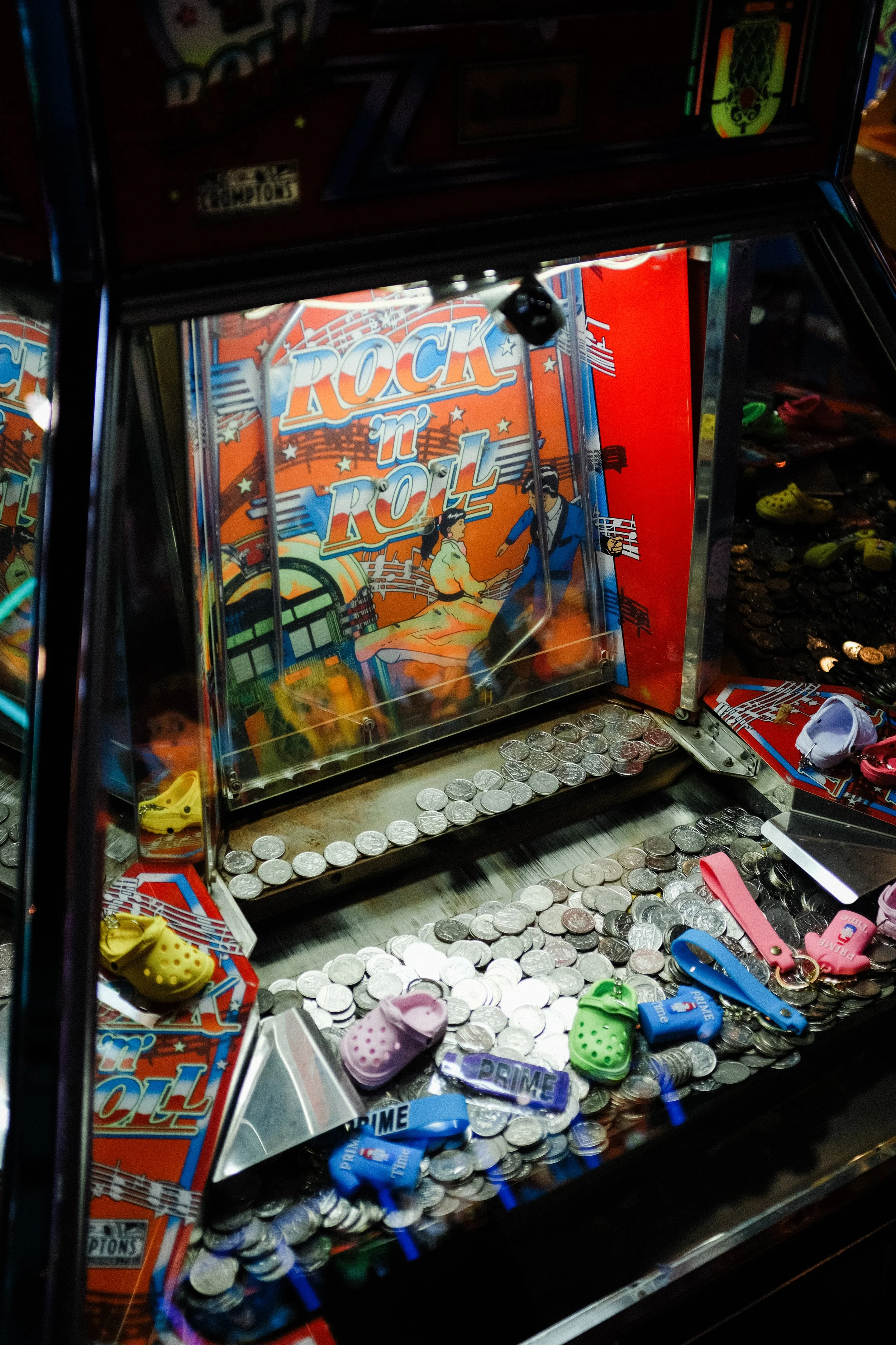 A vintage pinball machine named 'Rock n Roll' with a colorful backglass illustration of a man and woman dancing. The machine's playing field is filled with silver coins and small toy figurines, including colorful shoes and lanyards, surrounded by var