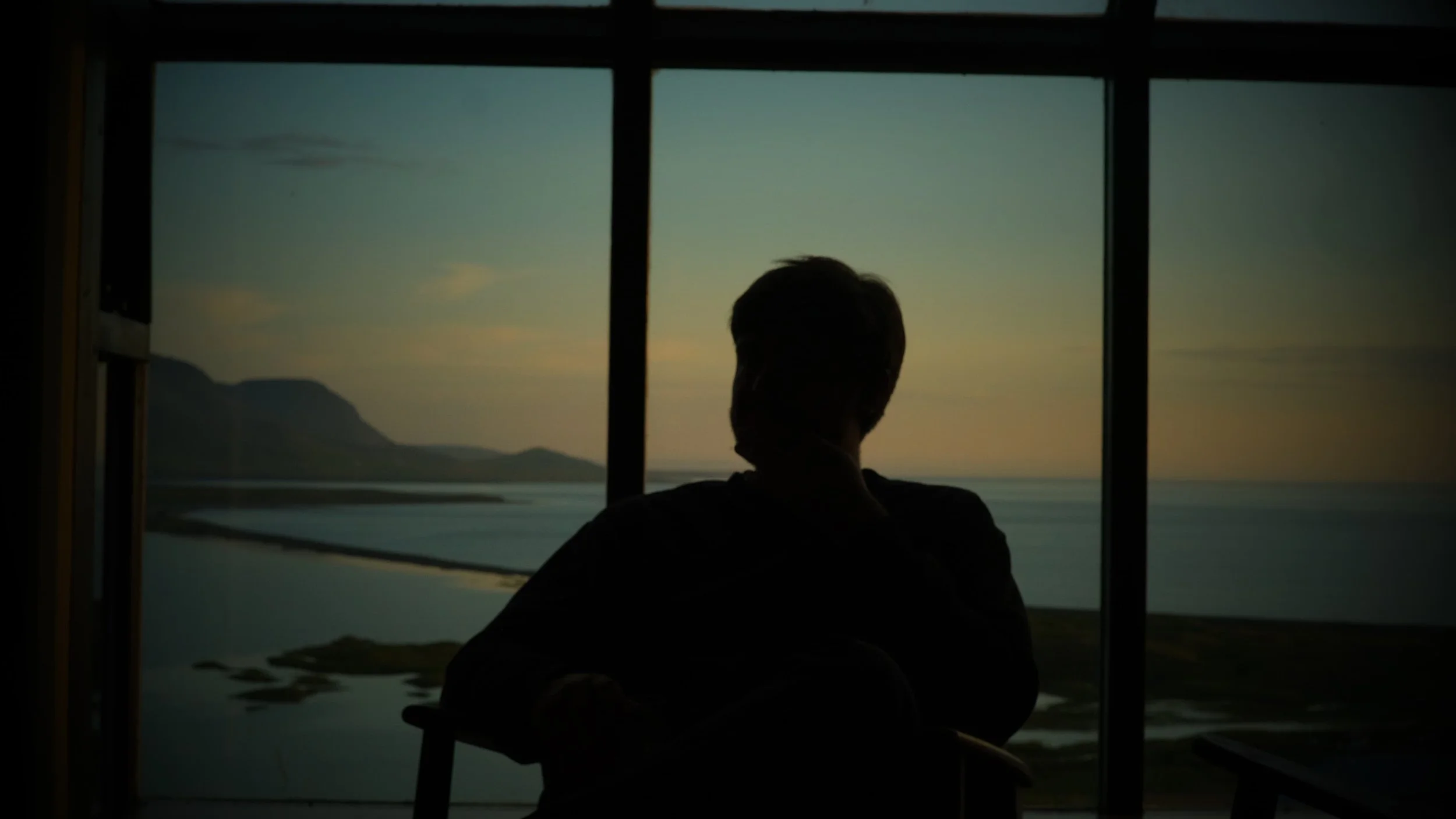 Person sitting by large window overlooking a lake and mountains at sunset.