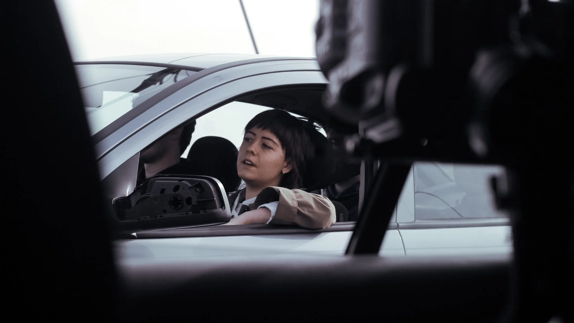 A woman with short dark hair and a beige jacket sitting in a car, seen through a camera or filming equipment.
