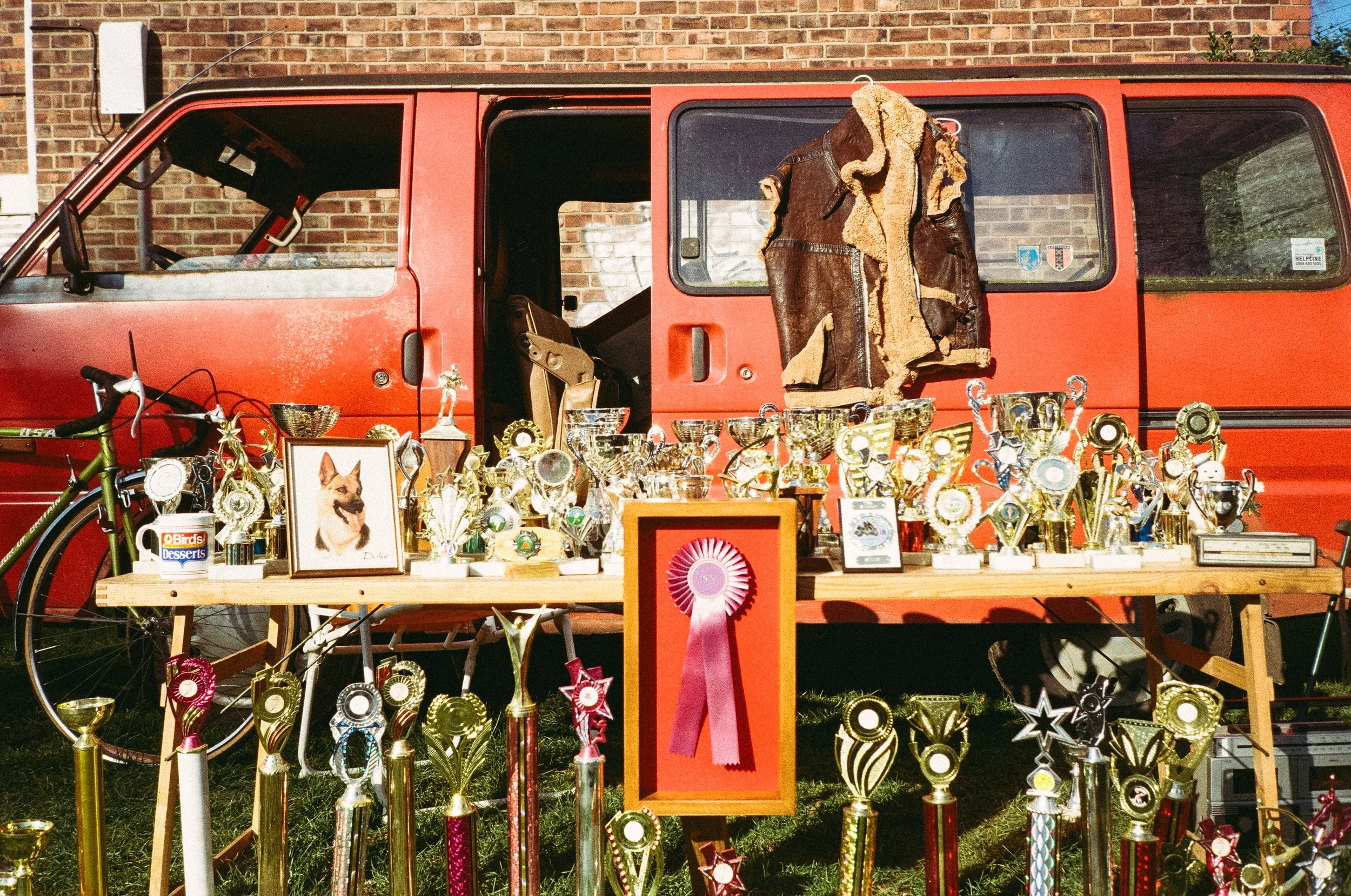 A sunset outdoor scene featuring a red van with trophies, awards, a framed dog picture, a pink ribbon, a bicycle, and a jacket hanging on the van, with a brick building in the background.