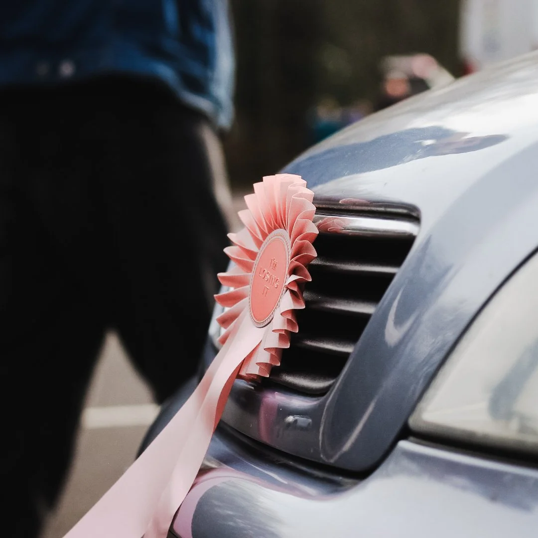 Close-up of a silver car's front grille with a pink and white rosette ribbon attached, indicating the car is lost.