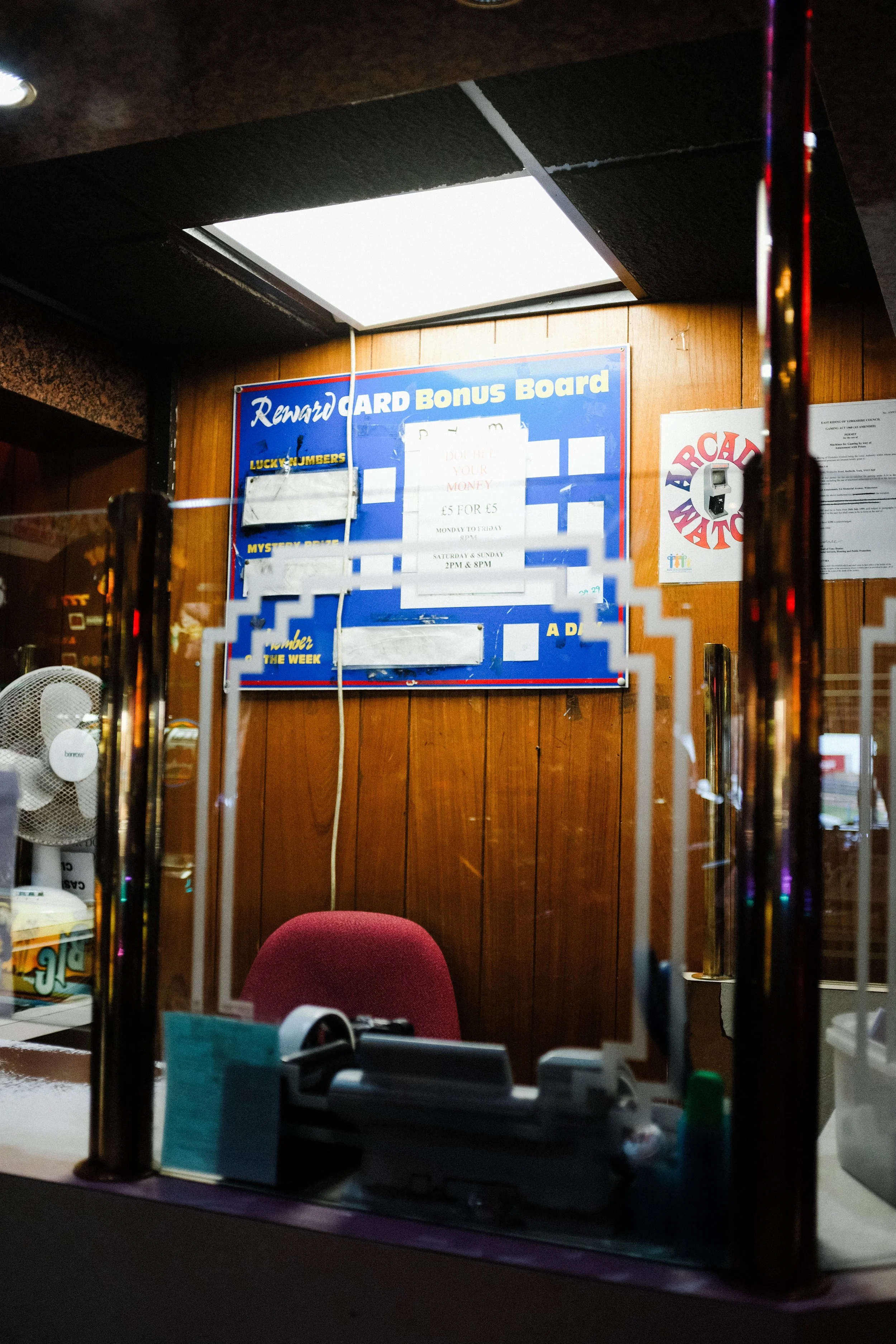 Inside view of a bar or pub with a red chair, a blue reward card bonus board on the wooden wall, and a red and white arcade match sign. The photo is taken through a window with reflections.