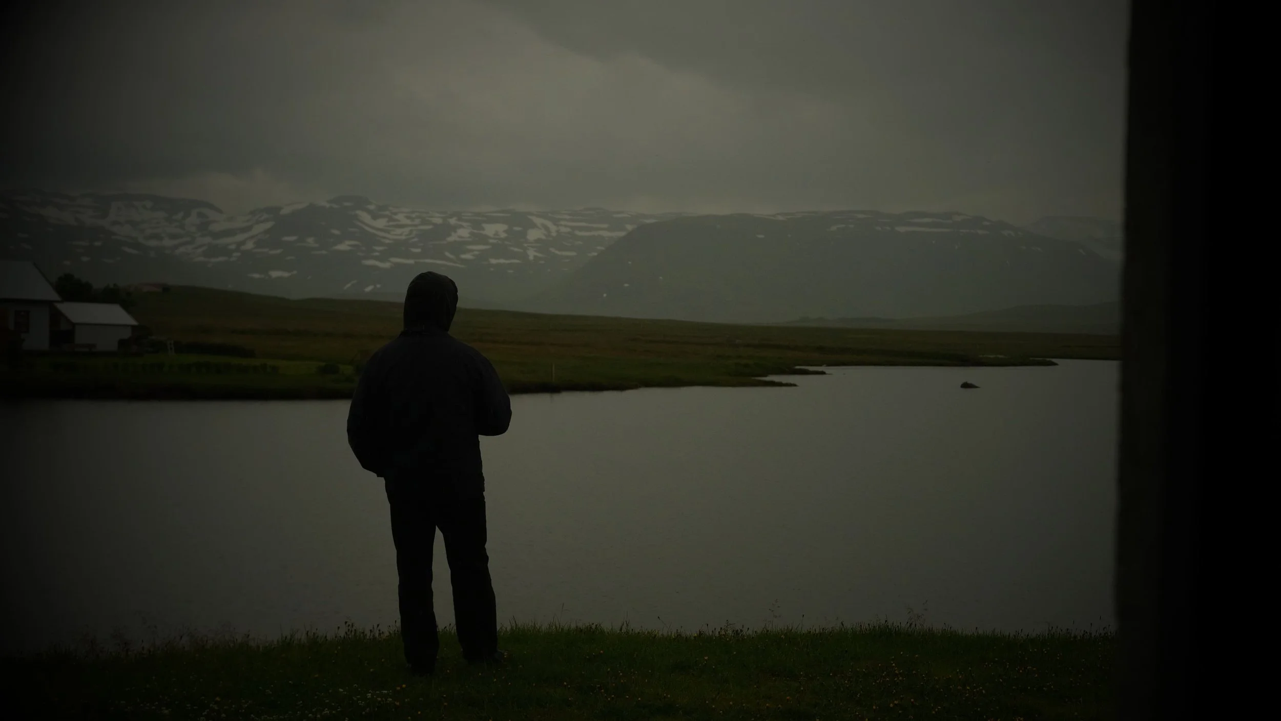 Person in dark clothing standing by a lake, looking at distant mountains with snow patches, under cloudy sky