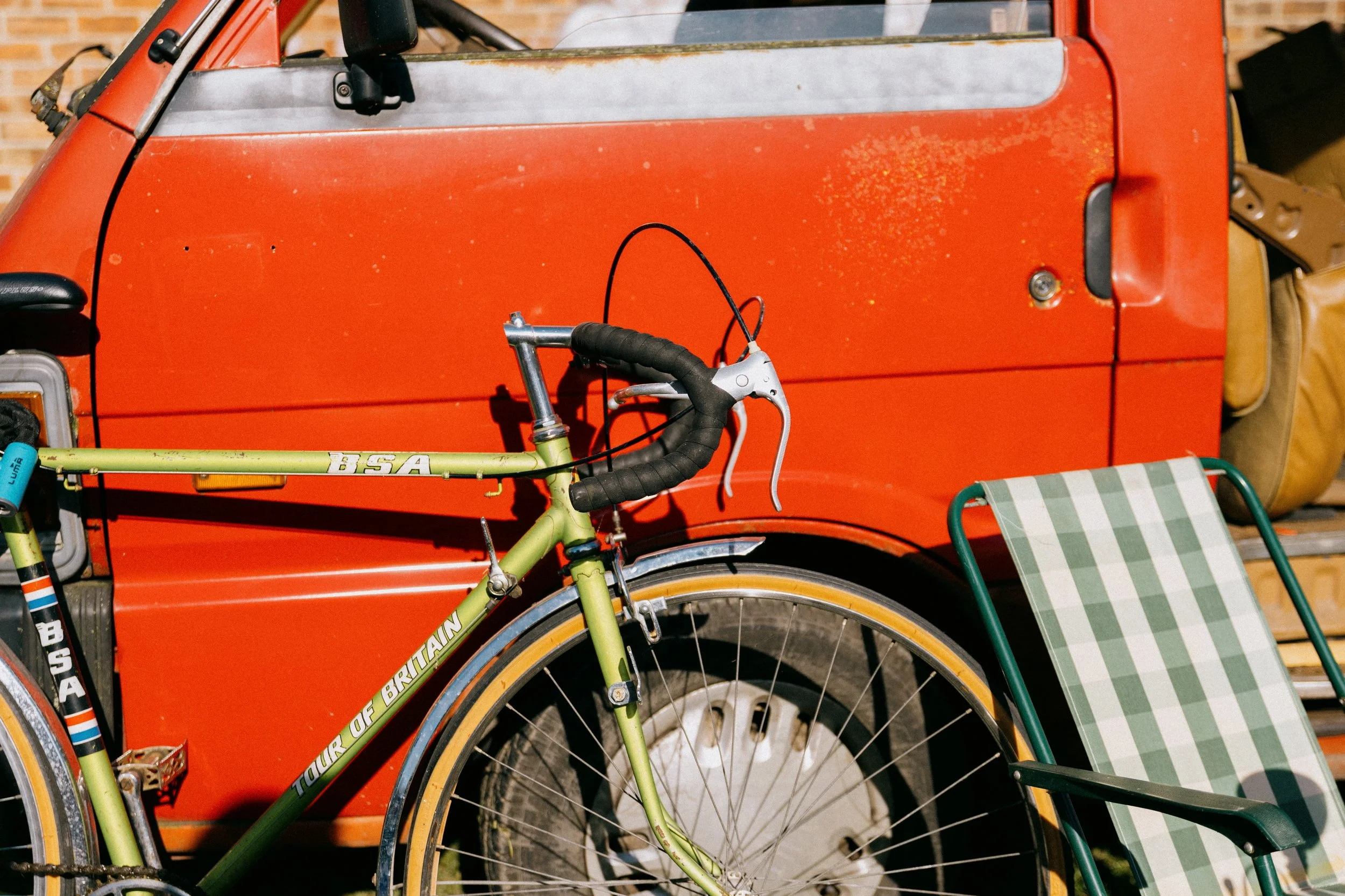 A green touring bicycle leaning against an orange vintage trailer, with a green and white checkered chair nearby.