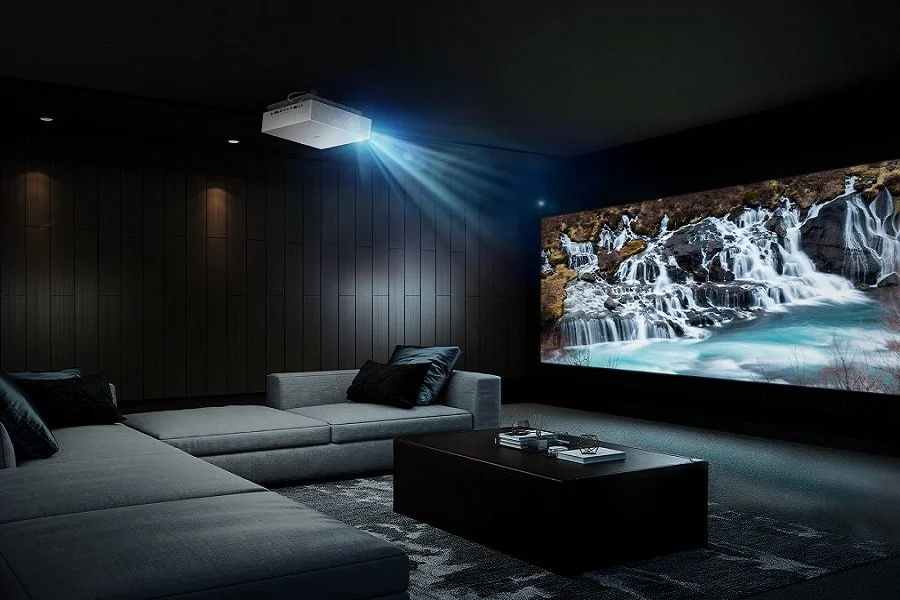 A modern home theater with a large screen displaying a waterfall scene, dark walls, gray sectional sofa with black pillows, and a ceiling-mounted projector projecting blue light.