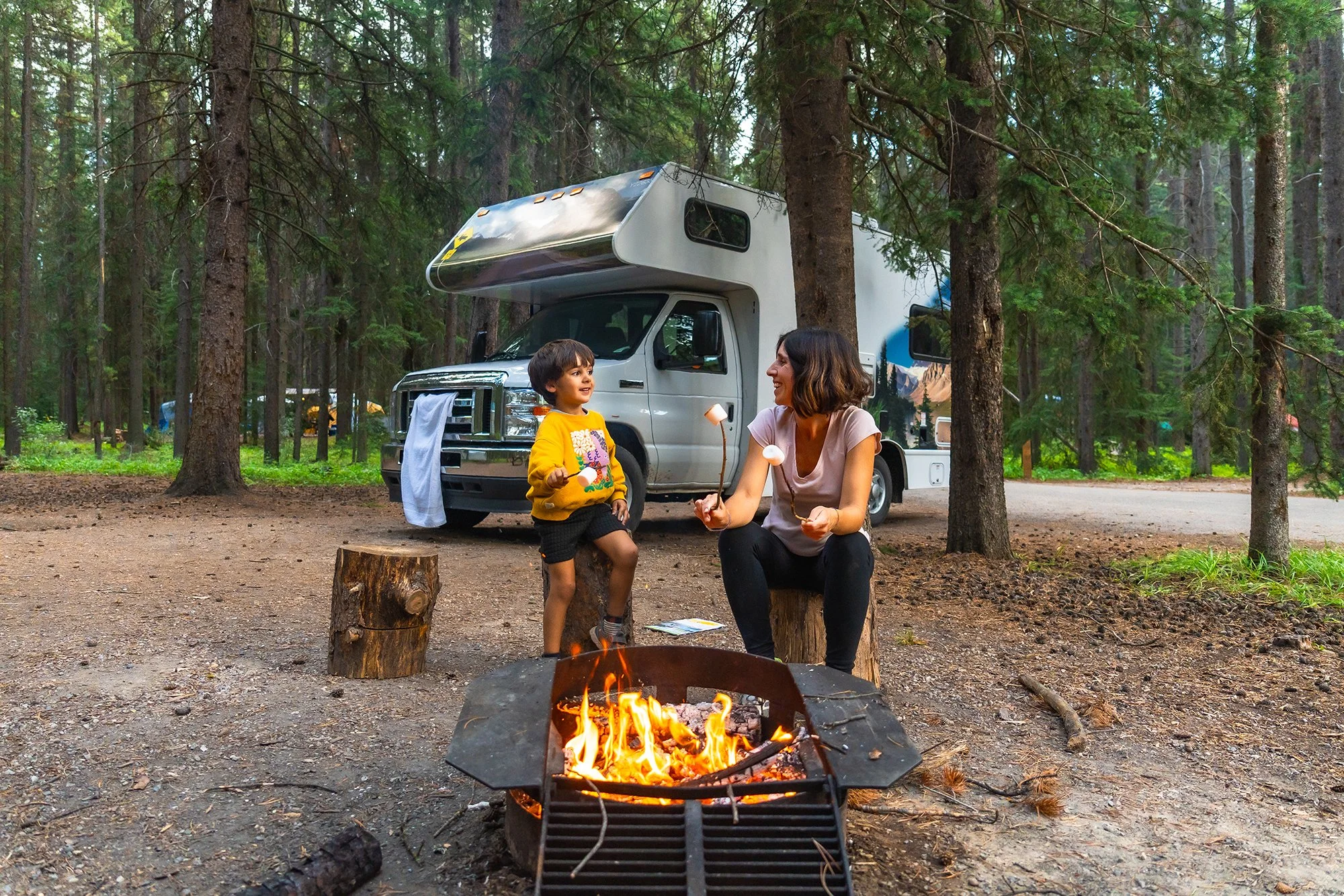 Woman and small child roasting marshmellows over camp fire with RV in the background in the woods
